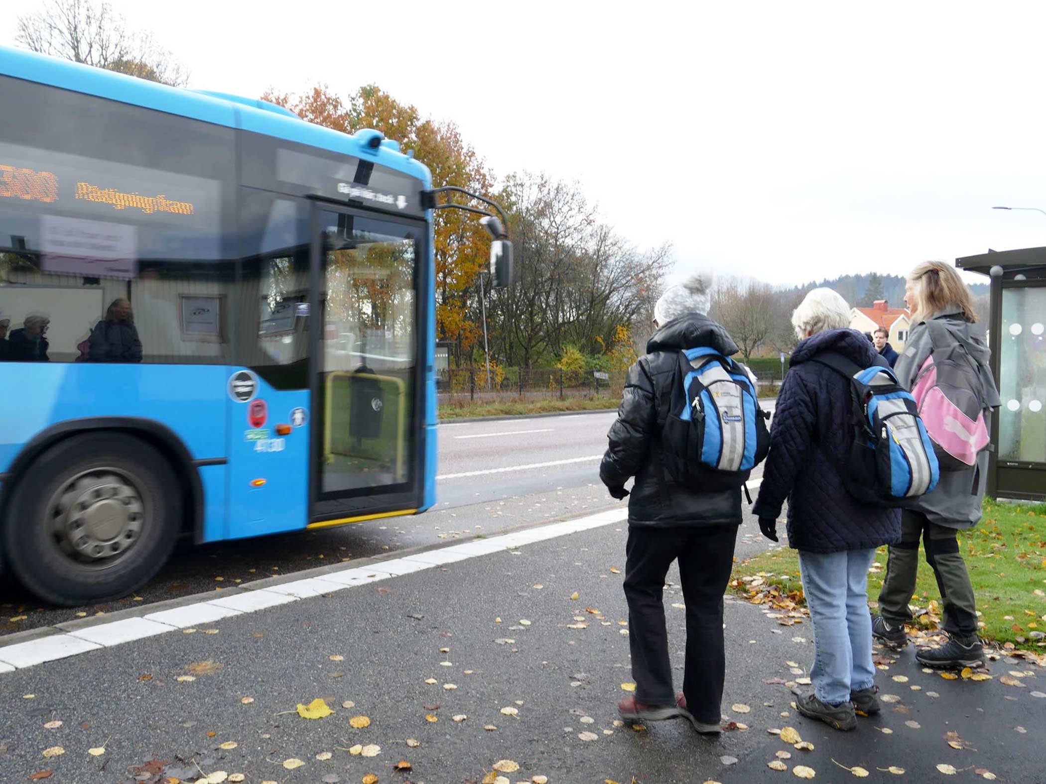 Tre personer väntar på buss vid hållplats en höstdag.