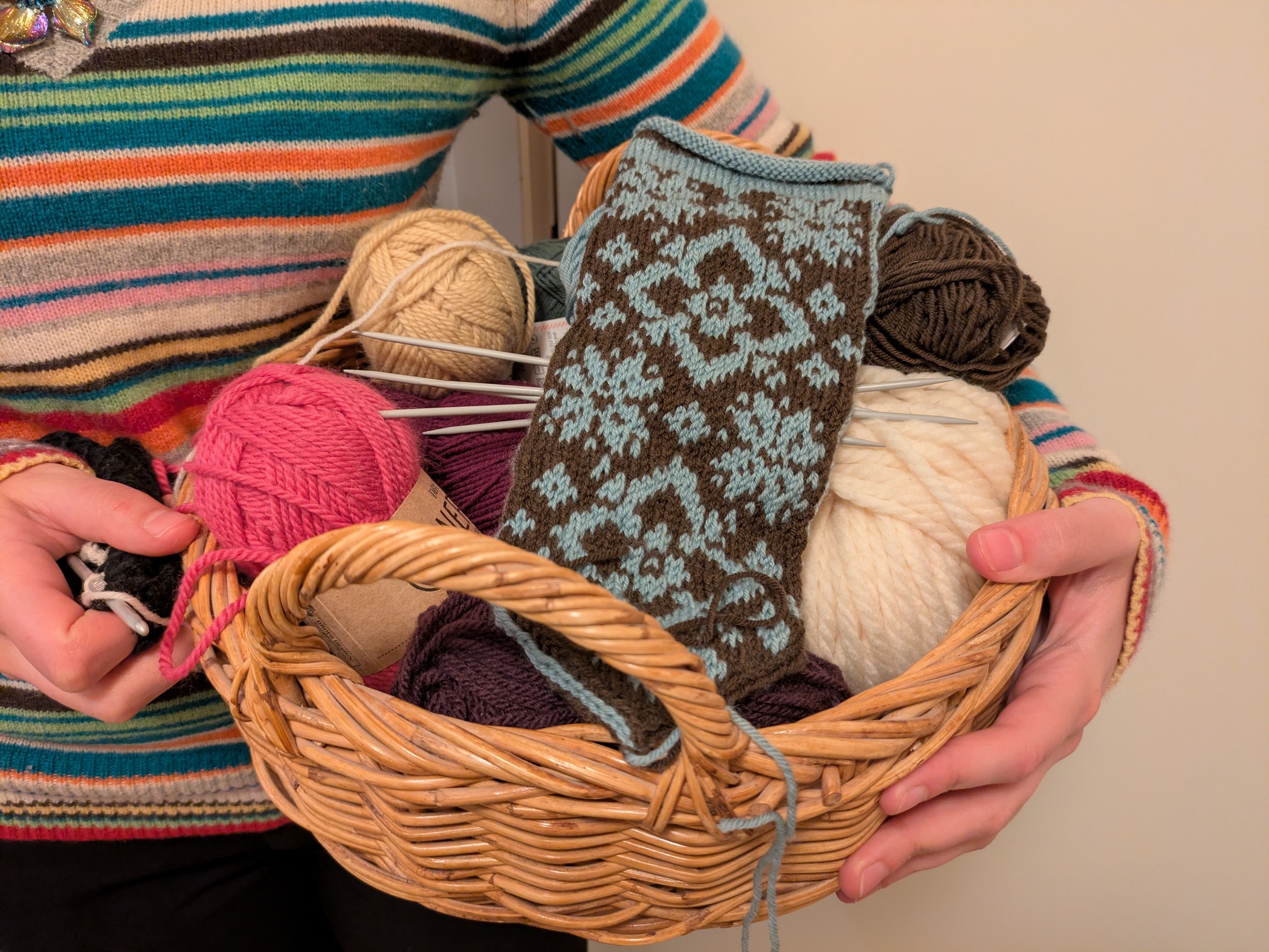 En person i färgglad tröja håller en korg fylld med garnnystan i olika färger och ett påbörjat stickprojekt i blått och brunt mönster syns.
