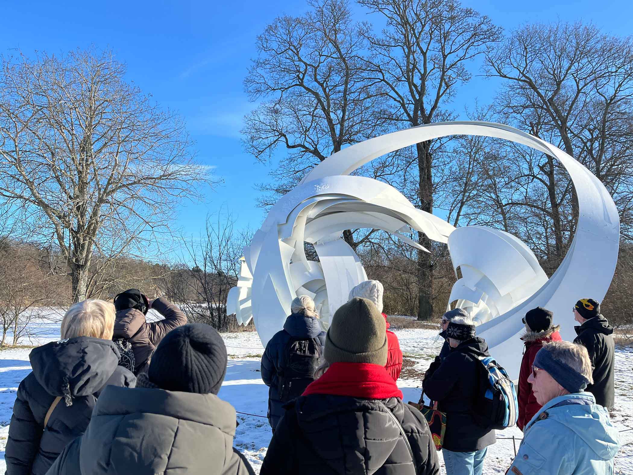 En grupp människor står samlade utomhus vintertid och lyssnar på en guide framför en stor vit abstrakt skulptur i ett snötäckt landskap.
