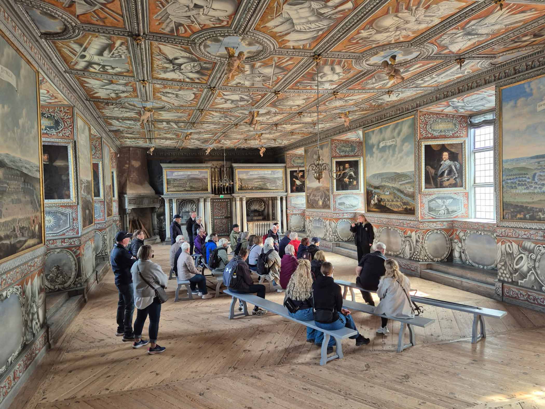 En grupp människor lyssnar på en guide i ett historiskt rum med målningar på väggar och tak.
