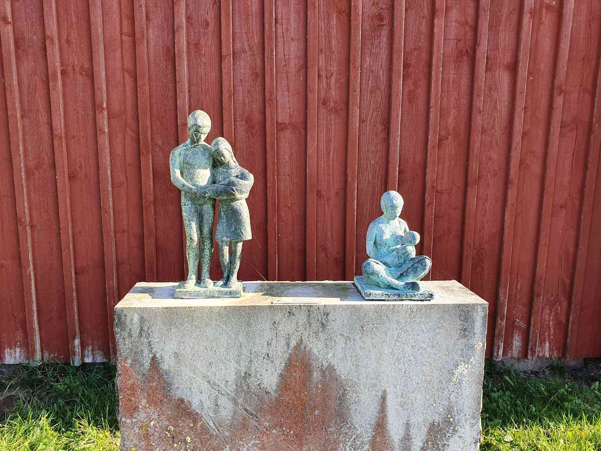 Två bronsstatyer på ett stenfundament framför en röd trävägg.