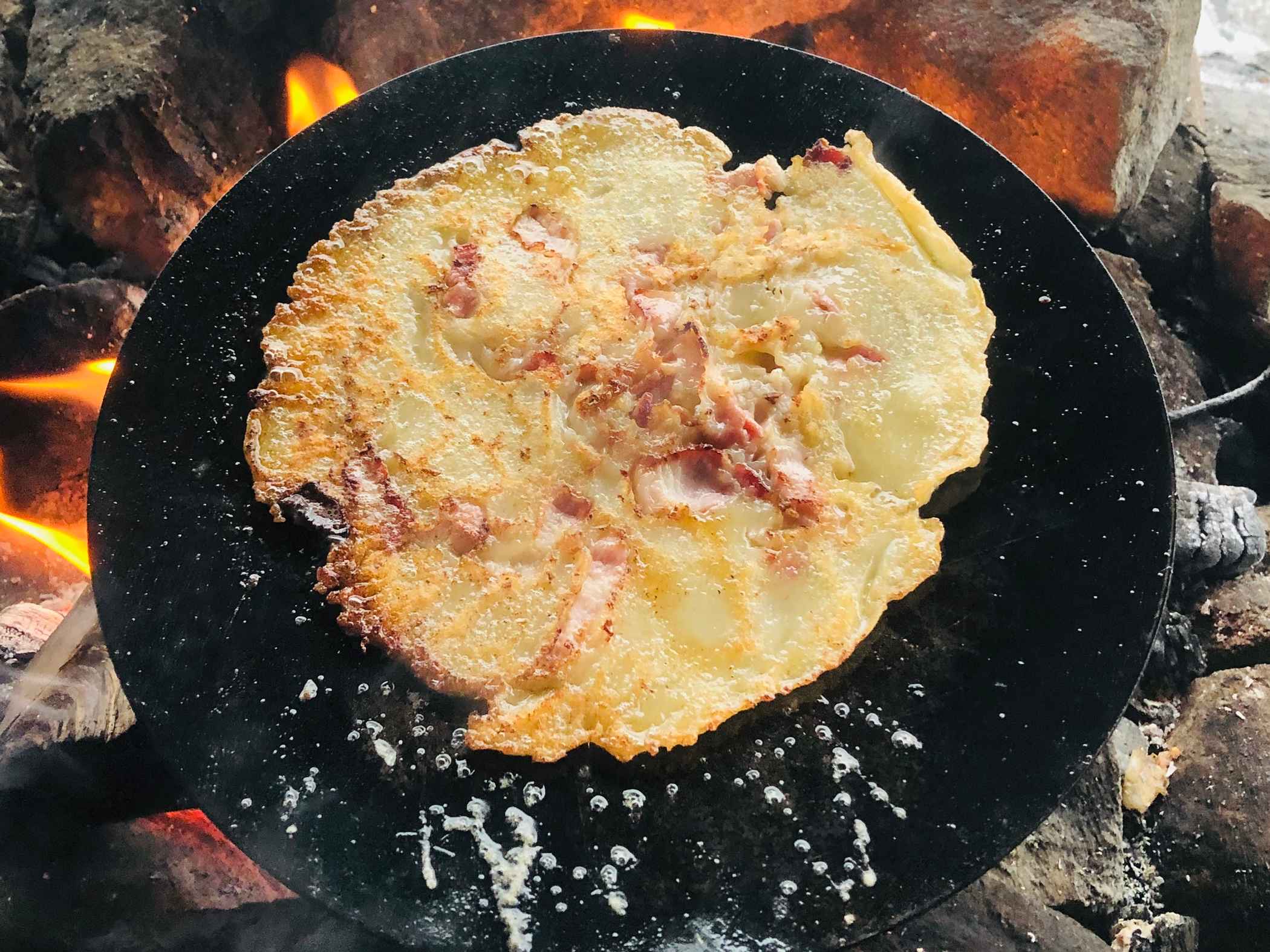 En raggmunk med fläsk steks över öppen eld på en stekhäll.