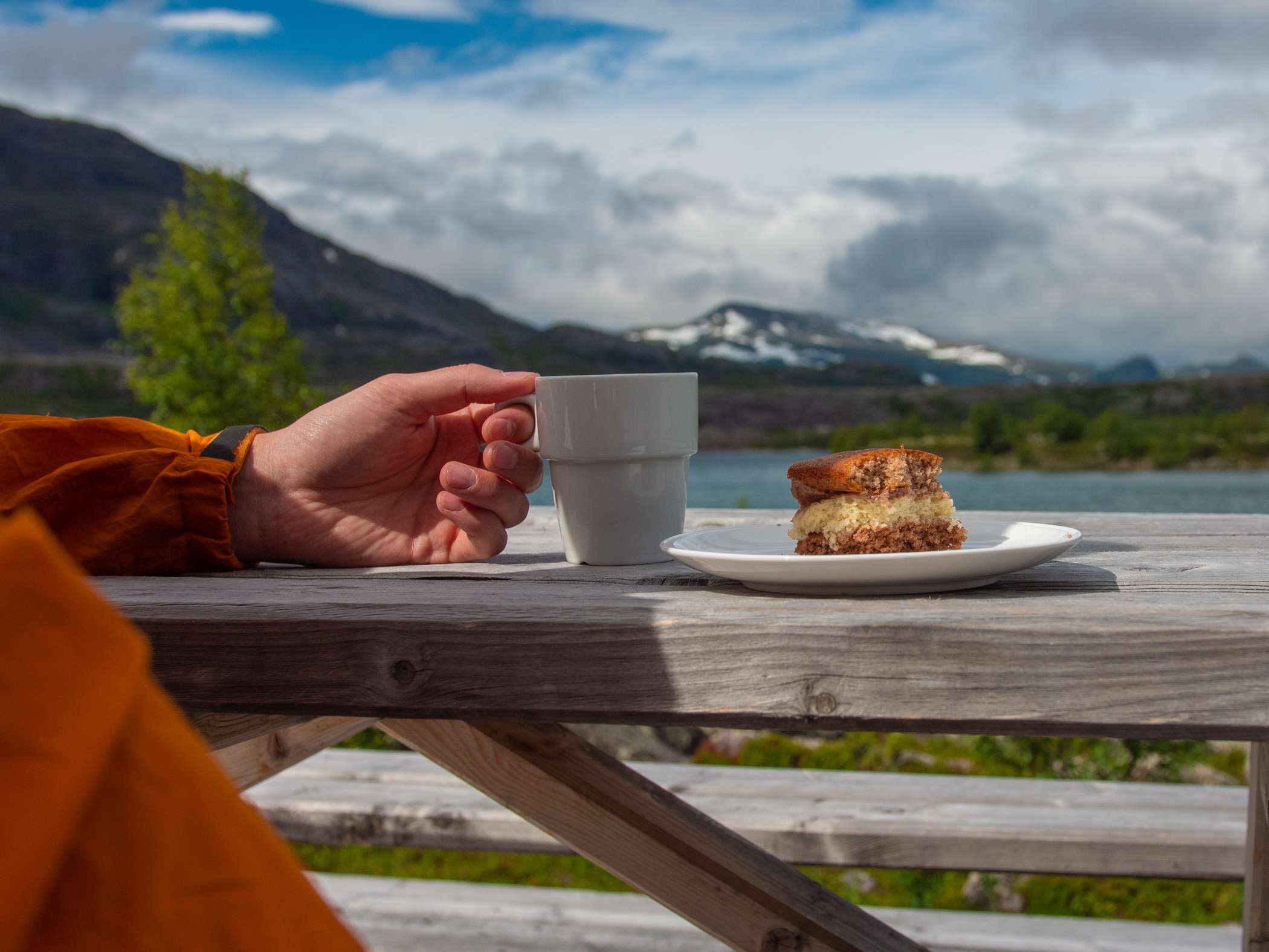 En person håller en kaffekopp bredvid ett fat med kaka och i bakgrunden syns utsikt över fjäll och sjö.
