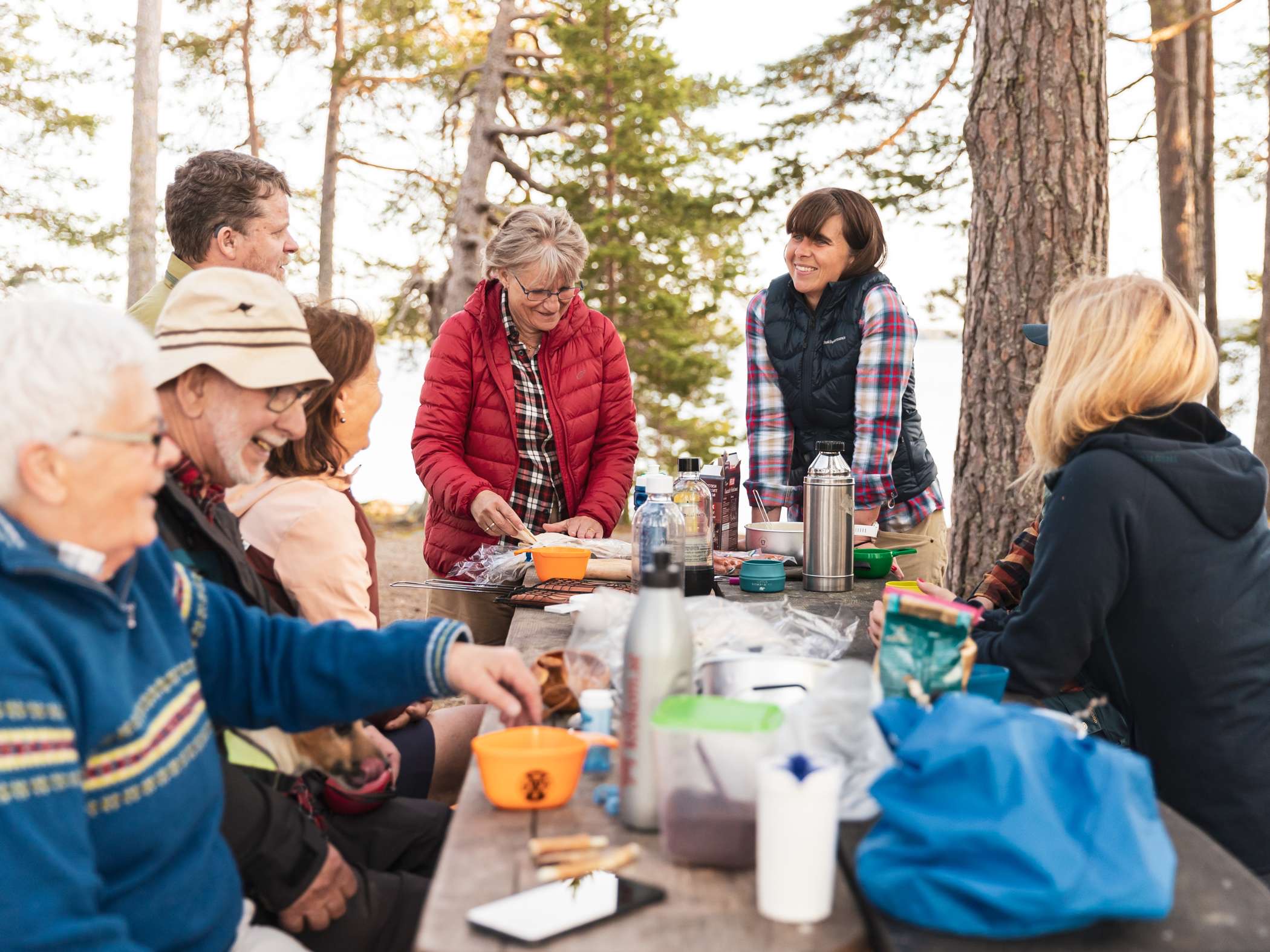 En grupp människor sitter runt ett bord i skogen och fikar tillsammans.