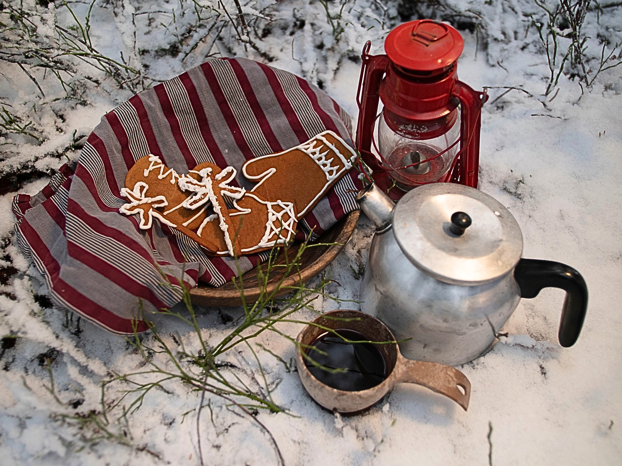 En bricka med pepparkakor, en kaffepanna och en röd lykta står på snötäckt mark, bredvid en kåsa med varm dryck.