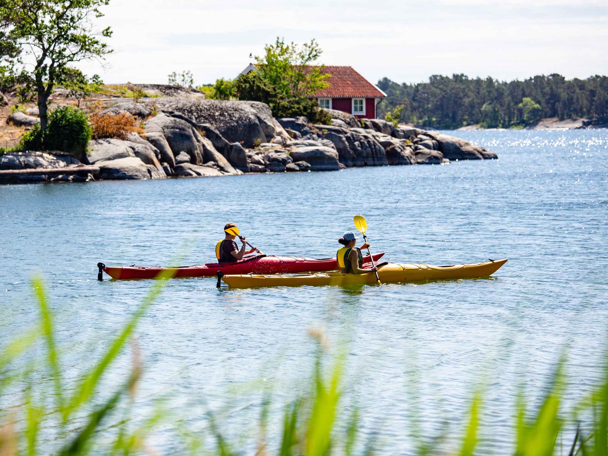 Två personer paddlar varsin kajak nära klippor och ett rött hus vid havet.