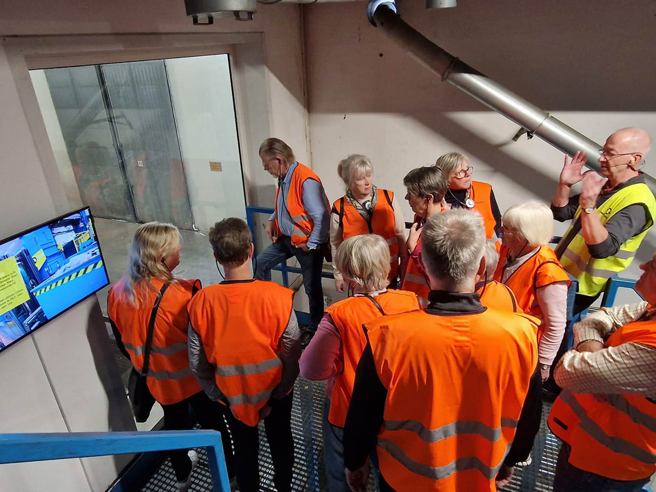 En grupp människor på studiebesök i en industri bär orangea västar och lyssnar på en guide.