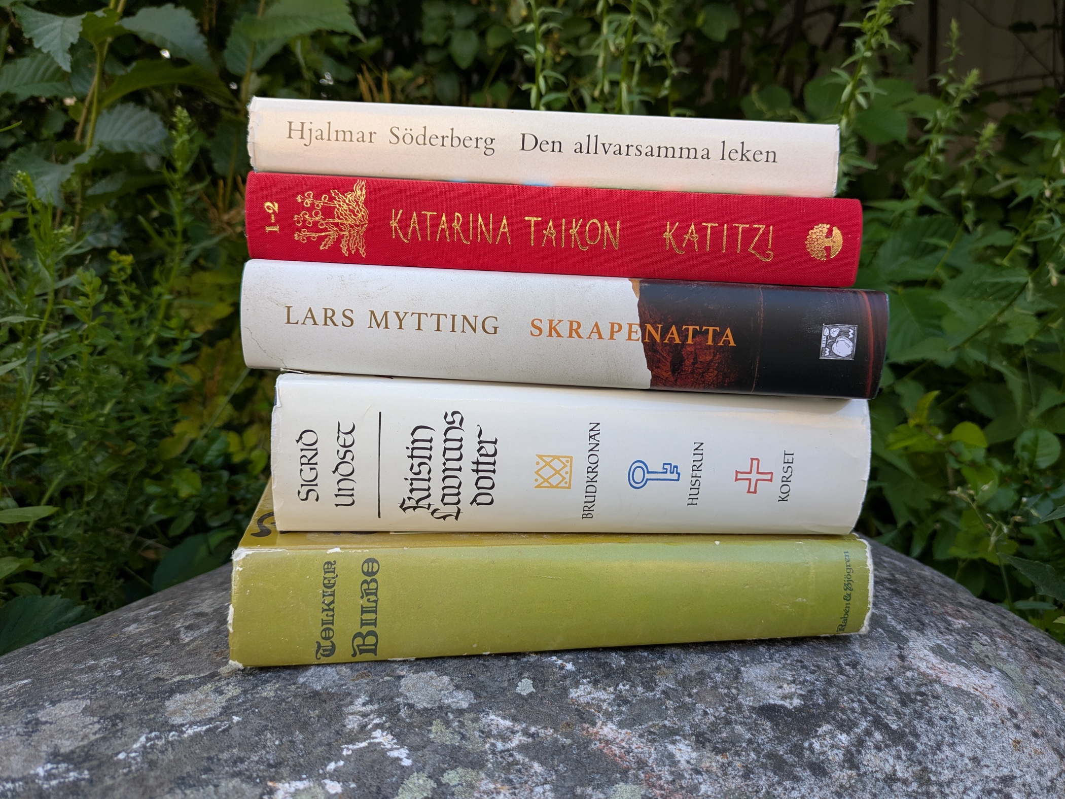 En hög med böcker ligger på en sten i en grön trädgård.