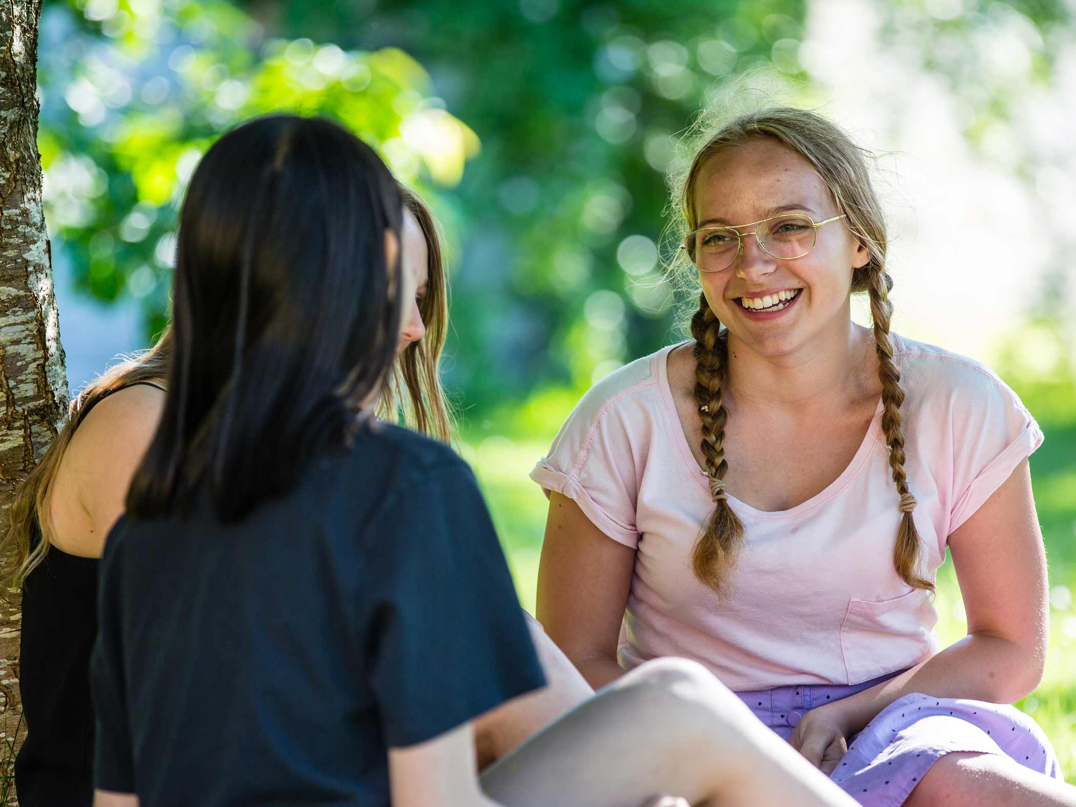 Två unga kvinnor sitter utomhus i grönskande miljö och samtalar.