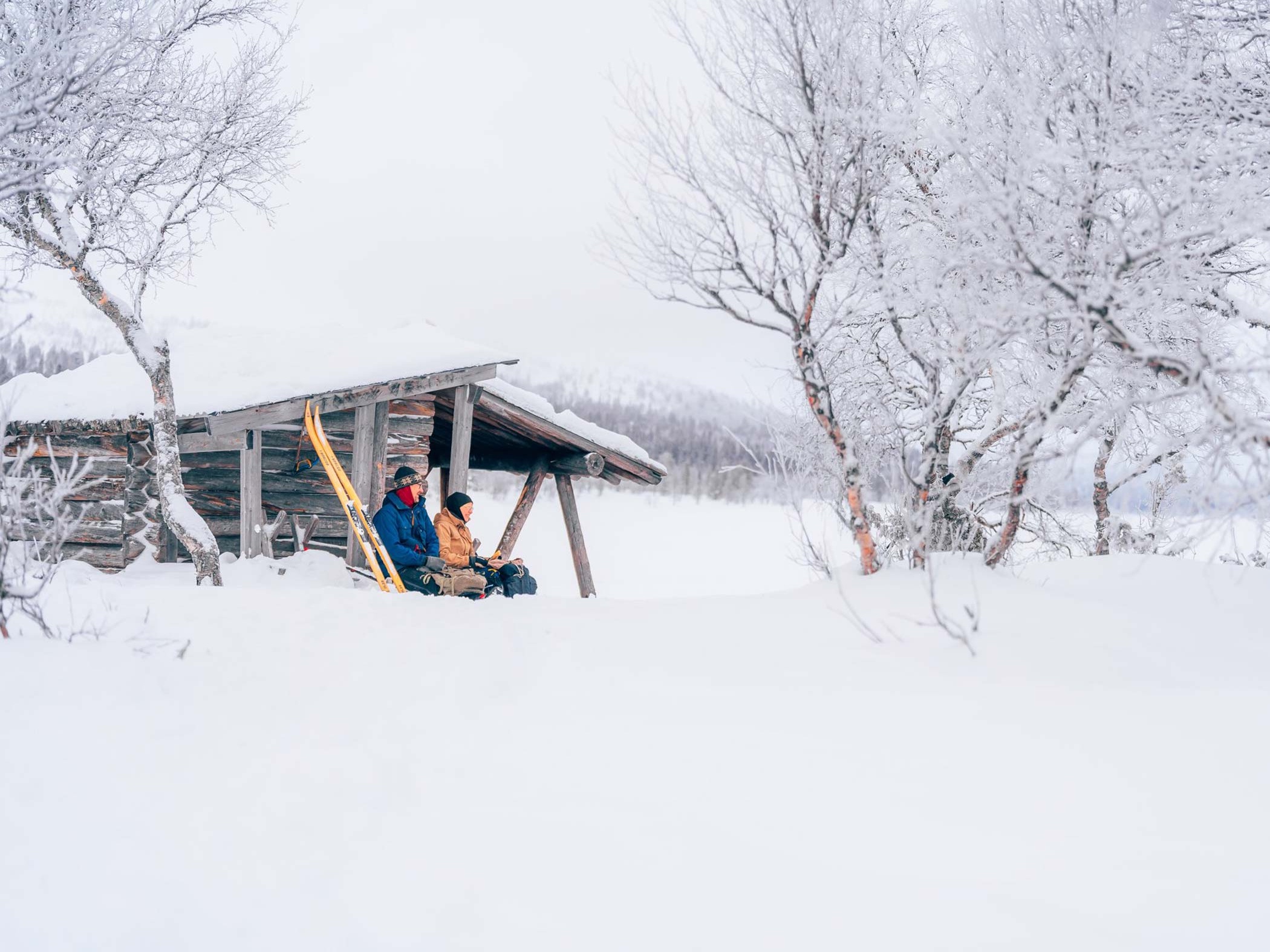 Två personer sitter vid ett vindskydd i ett snötäckt fjällandskap, med längdskidor lutade mot väggen och frostklädda träd runt omkring sig.