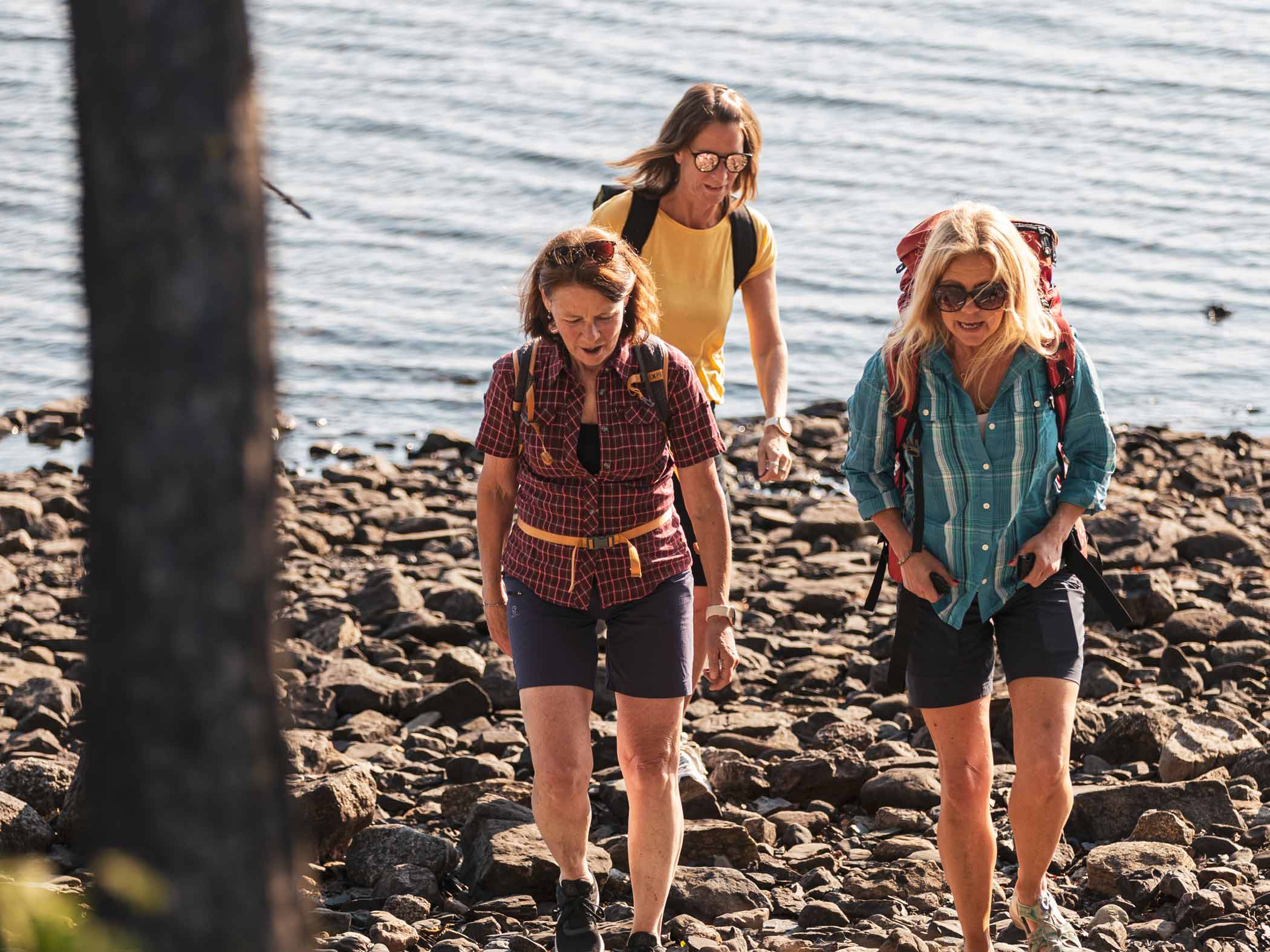 Tre kvinnor vandrar längs stenig strand vid vatten en sommardag.