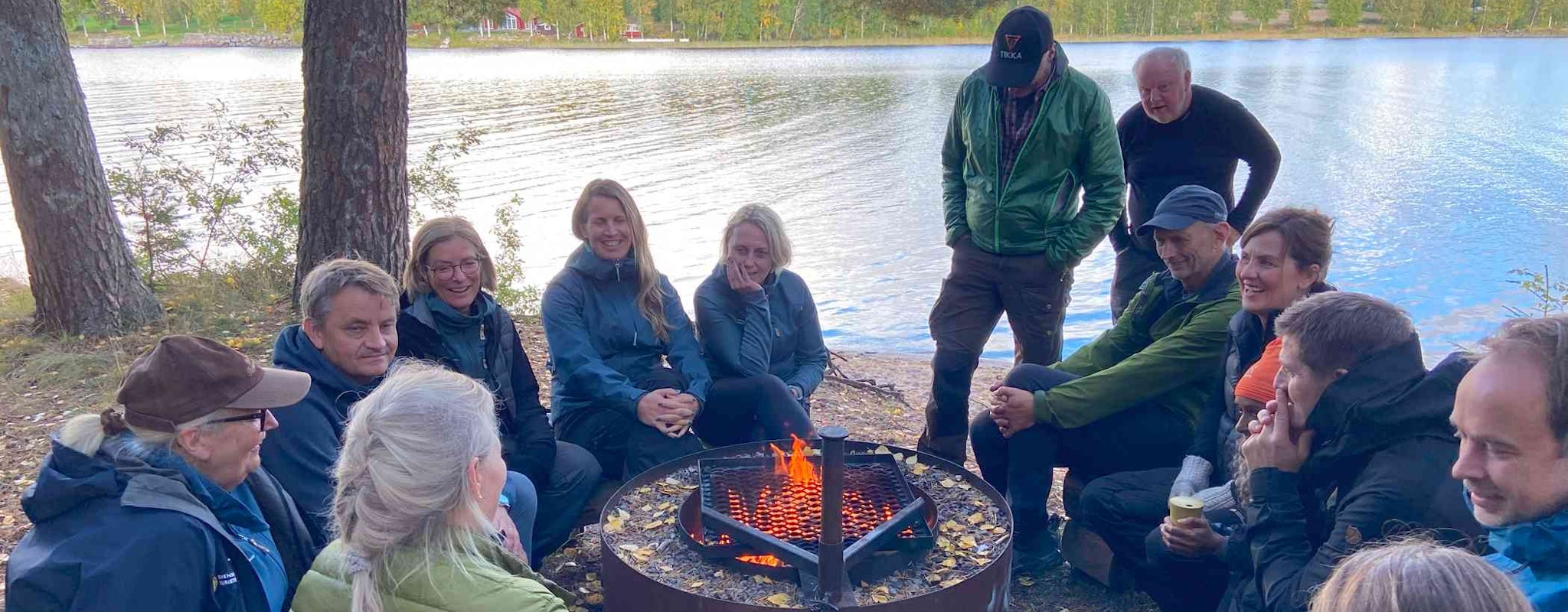En grupp människor sitter runt en eld vid en sjö och pratar under en paus på vandring.