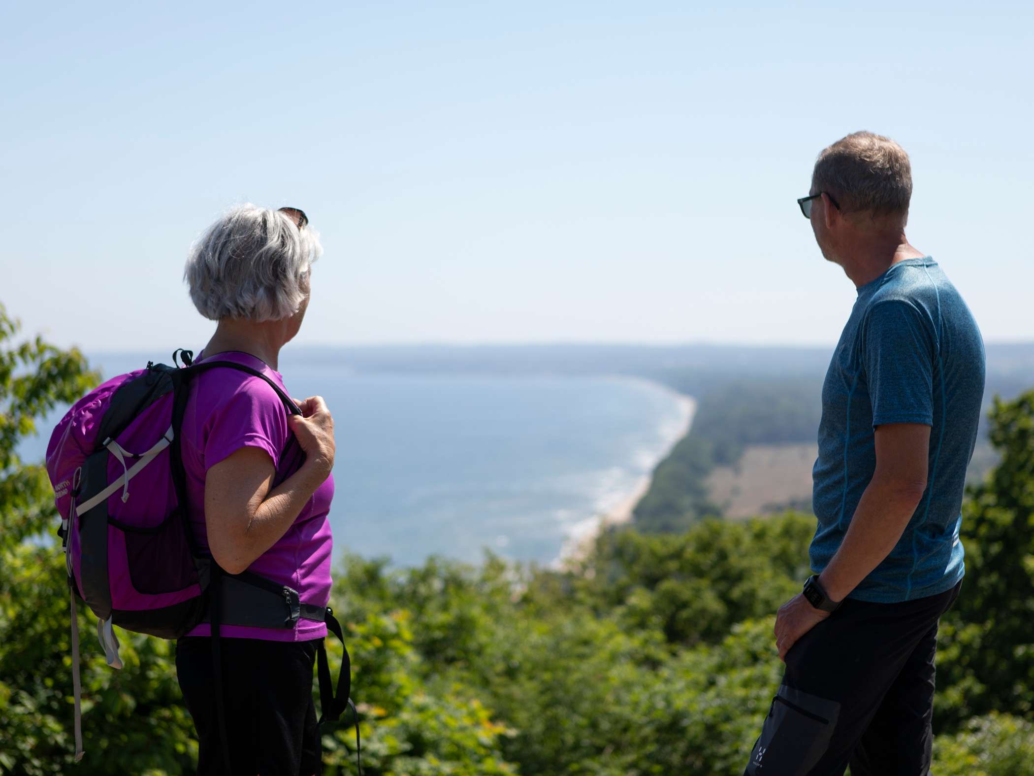 Två personer står på en höjd och blickar ut över havet och en lång kustlinje i solljus.