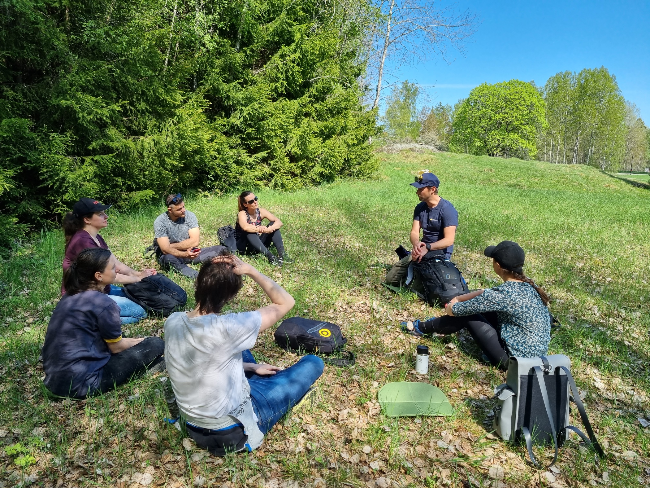 En grupp personer sitter i gräset i en glänta i skogen och vilar och pratar tillsammans.