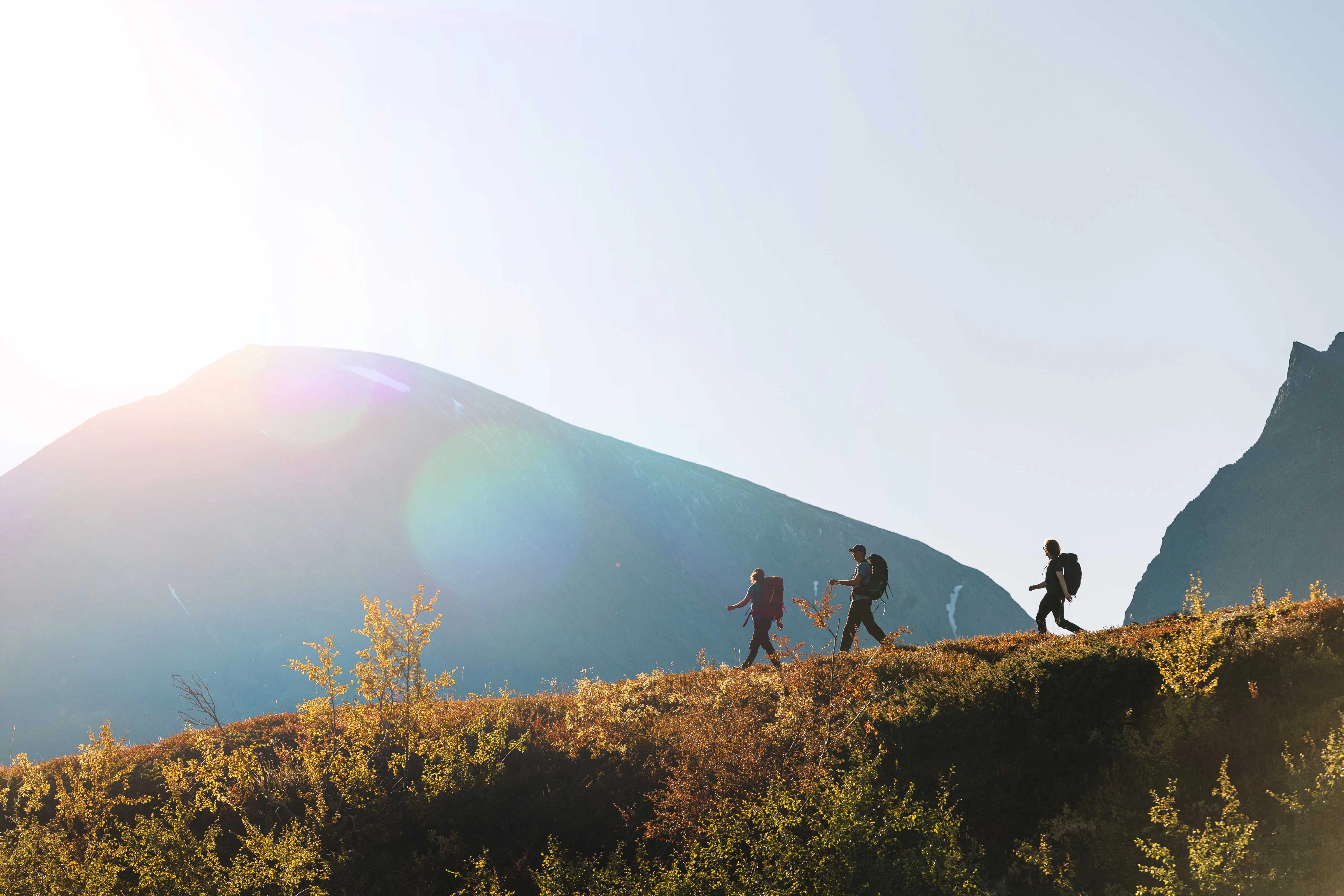 Tre vandrare går på en solbelyst fjällrygg med berg i bakgrunden.