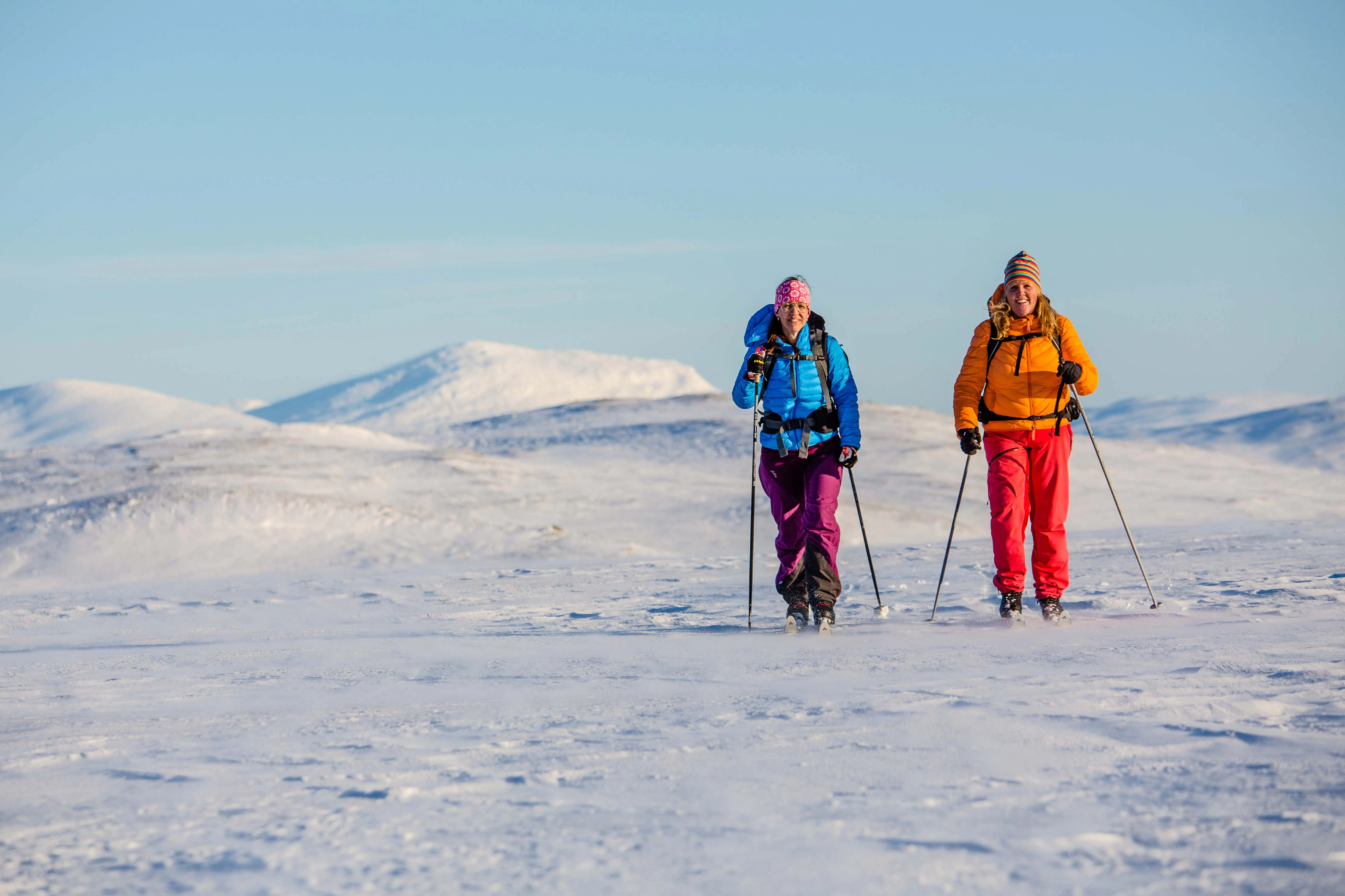 Två kvinnor åker längdskidor över ett öppet, snötäckt fjällandskap.
