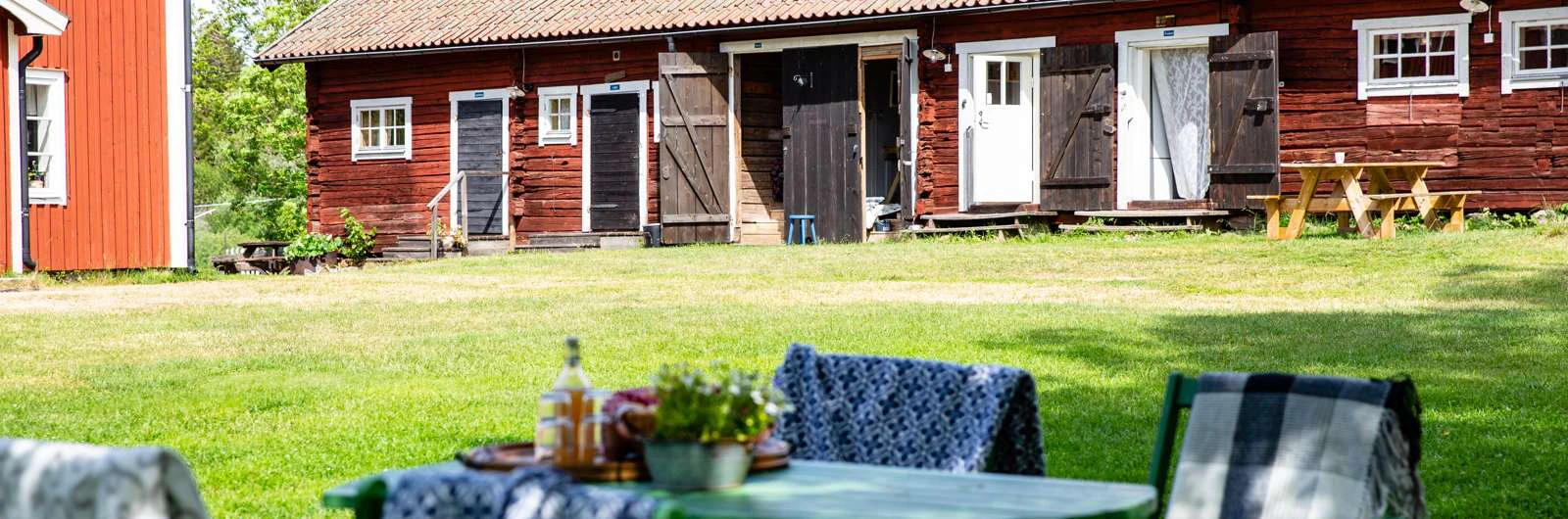 Ett grönskande gårdsplan med röda träbyggnader och vita dörrar i bakgrunden och ett dukat bord med dryck och filtar i förgrunden.