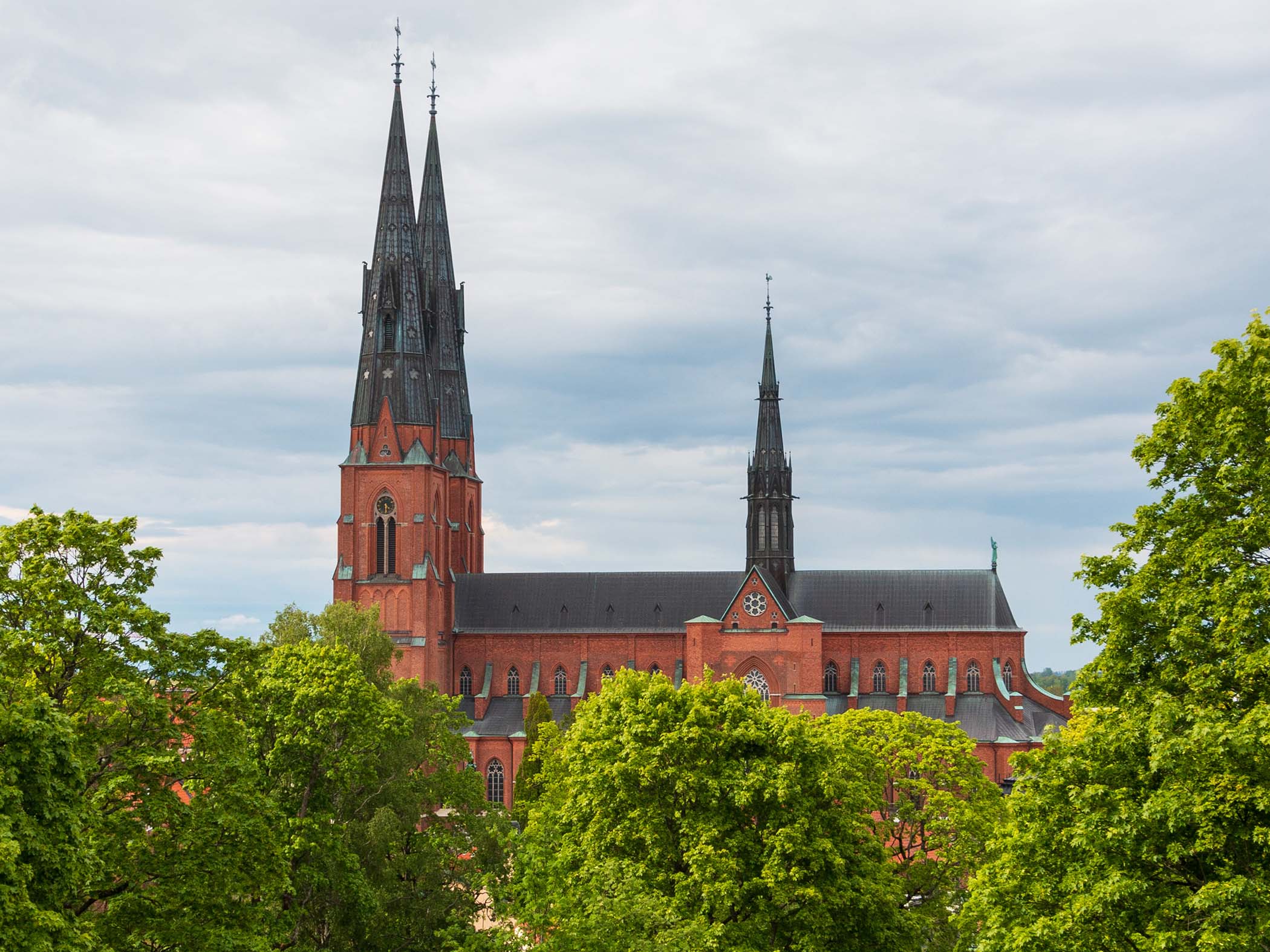 Uppsala katedral med två höga spiror, omgiven av grönskande träd under en molnig himmel.