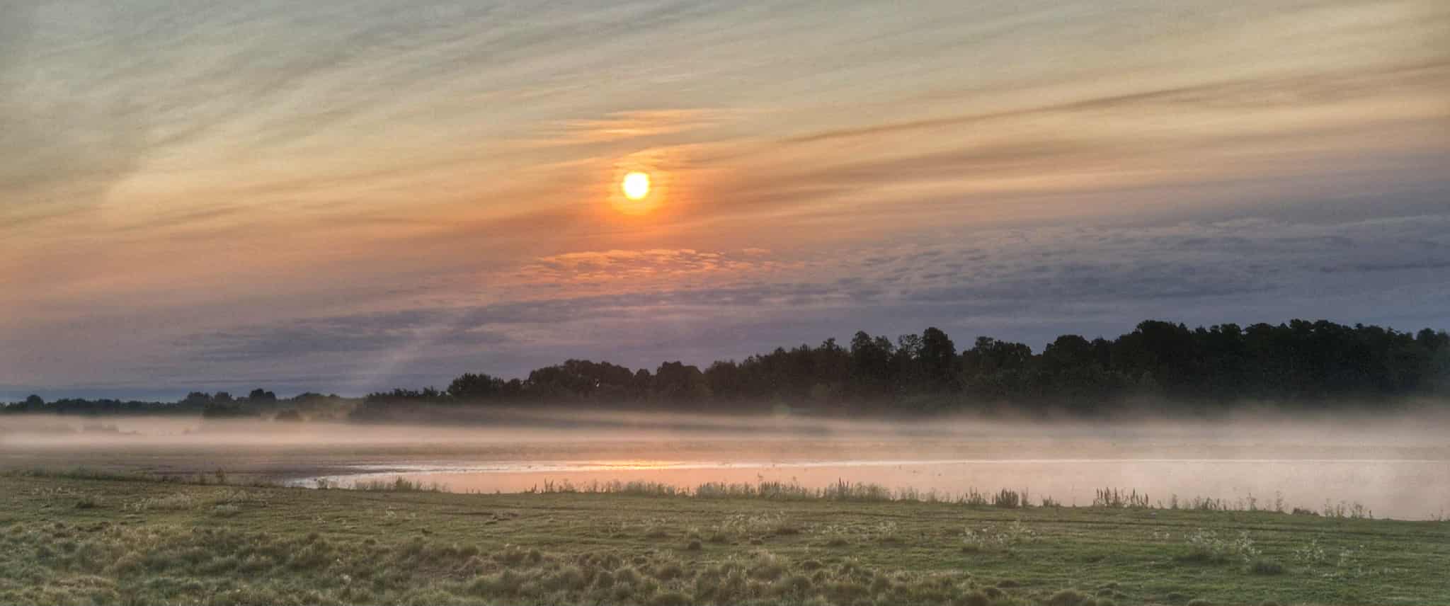 En dimmig morgon vid en sjö i Närke Bergslagen med solen som stiger över horisonten och färgar himlen i varma toner.