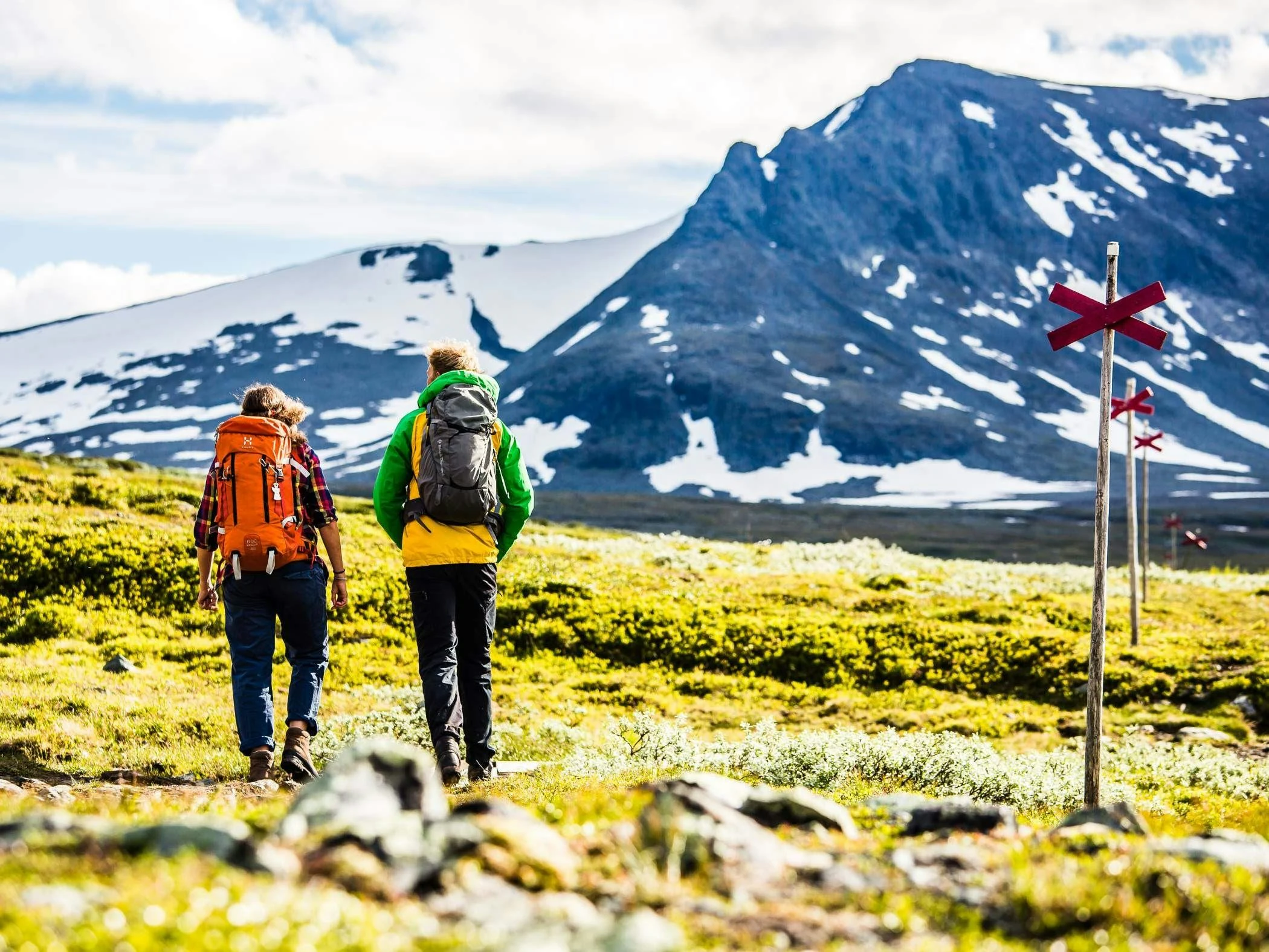 Två vandrare med ryggsäckar som följer markerad led genom fjällandskap med snöklädda berg i bakgrunden