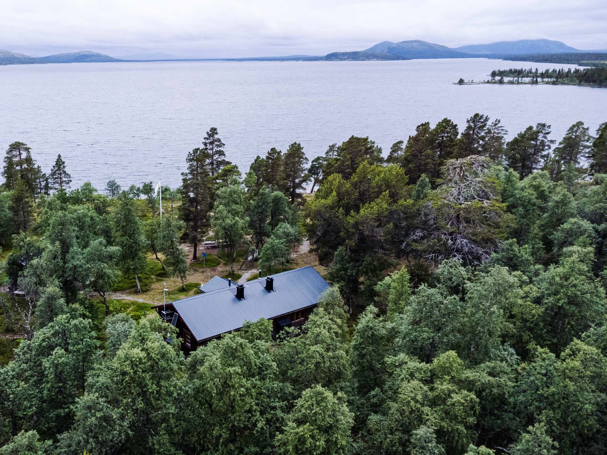 Flygbild av en stuga omgiven av skog, nära en sjö med berg i bakgrunden.