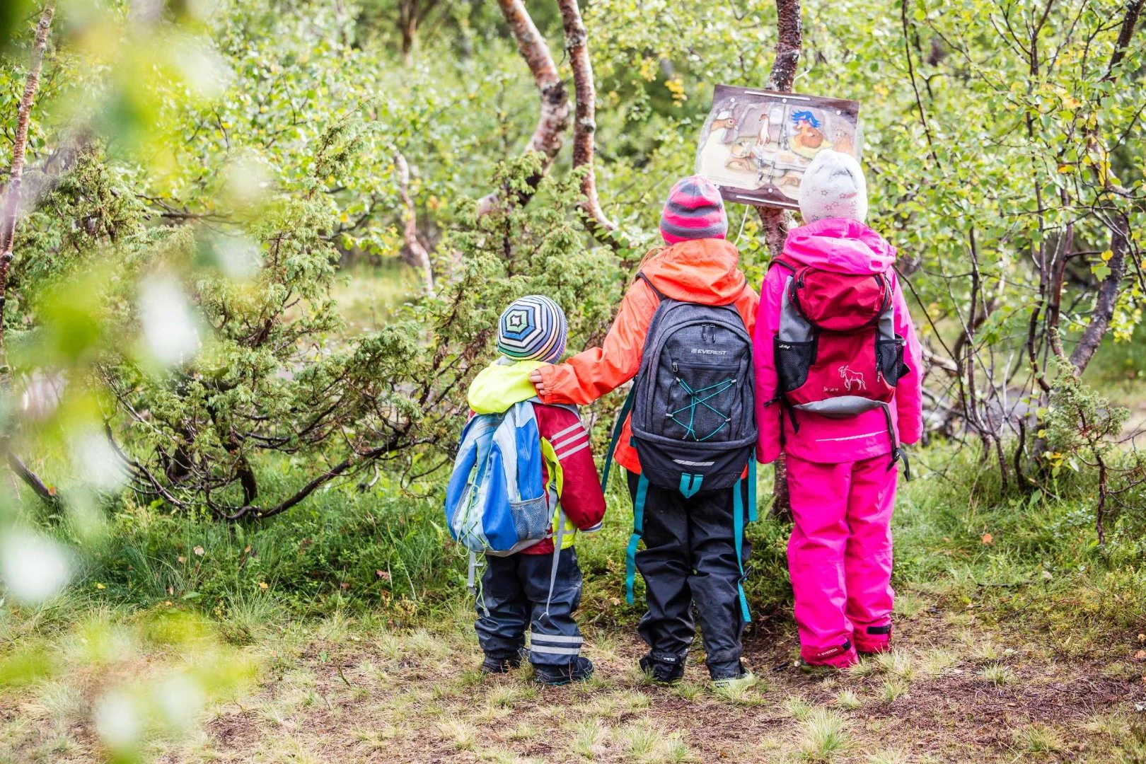 Tre barn med ryggsäckar står vid en skylt i grönskande fjällskog under en vandring.