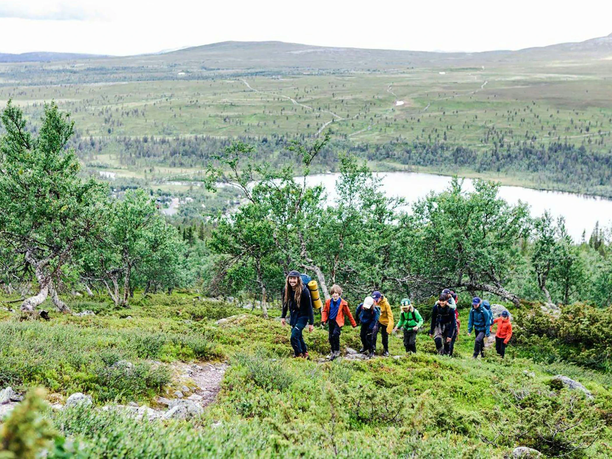 En grupp människor vandrar uppför en grönskande sluttning med utsikt över en sjö och fjäll i bakgrunden.