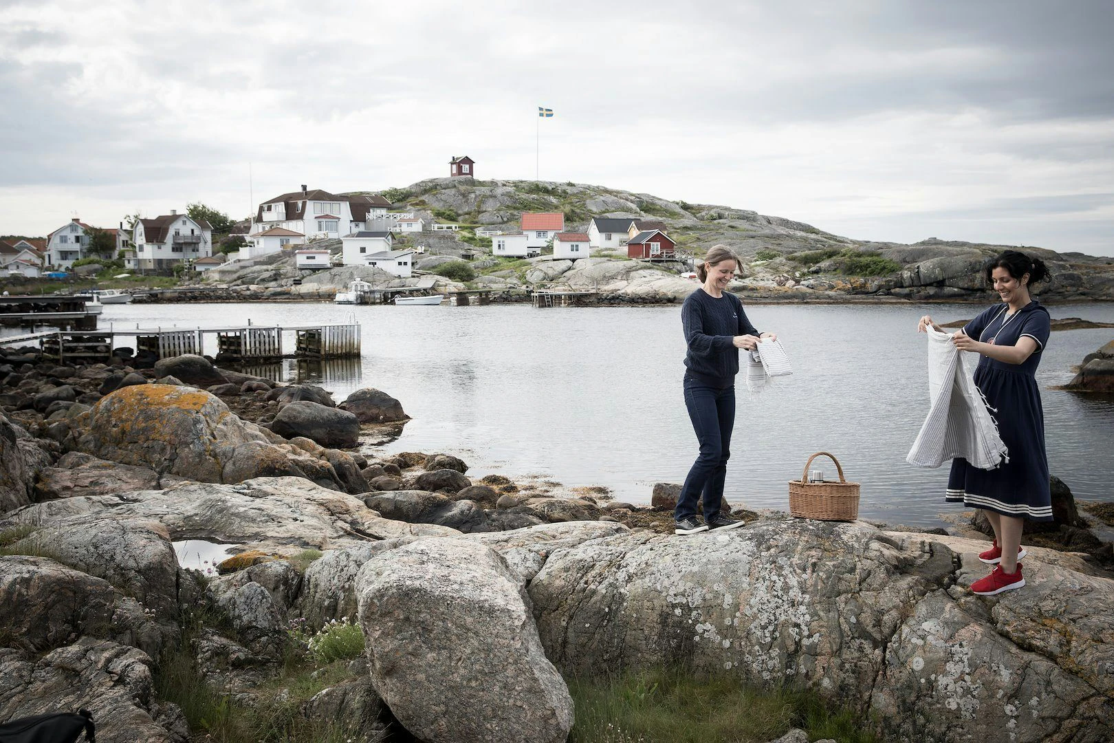 Två kvinnor dukar upp för picknick på klipporna vid havet, med en pittoresk kustby i bakgrunden.