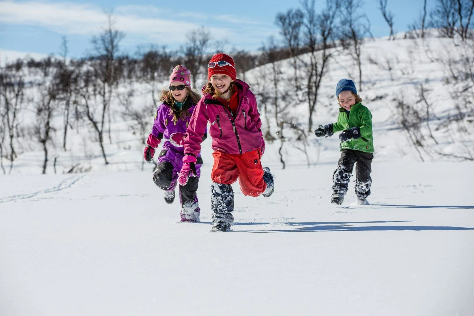 Tre barn leker och springer glada i snön en solig vinterdag i fjällmiljö.