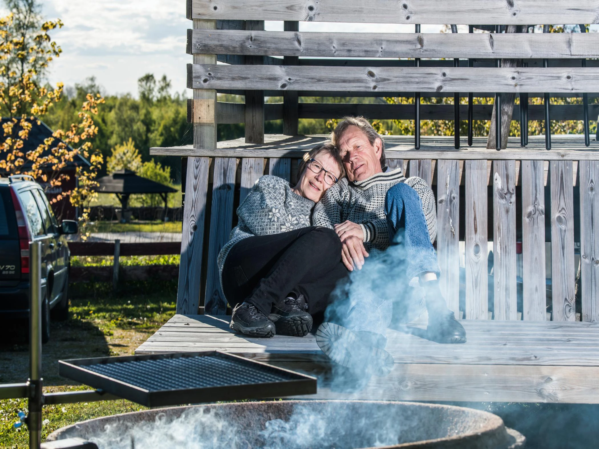 Par i ulltröjor sitter tätt tillsammans vid en träbänk intill en eldplats utomhus.