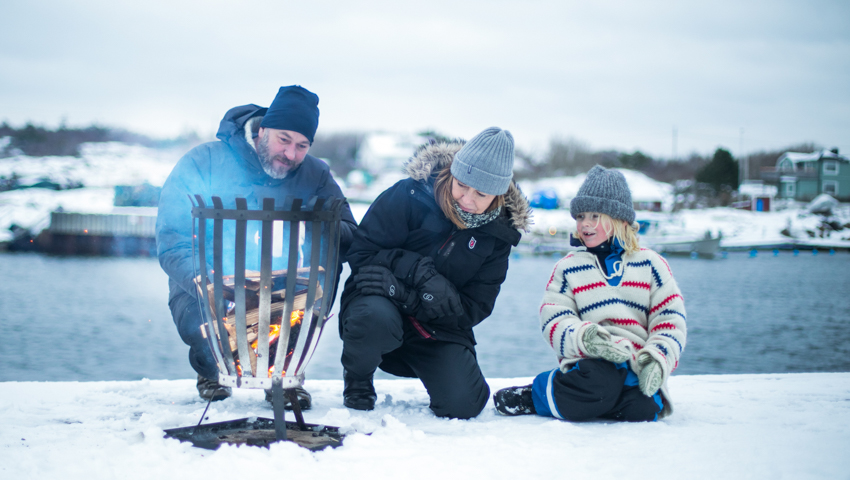 Familj eldar ved i ett vinterlandskap med vatten bakom sig.