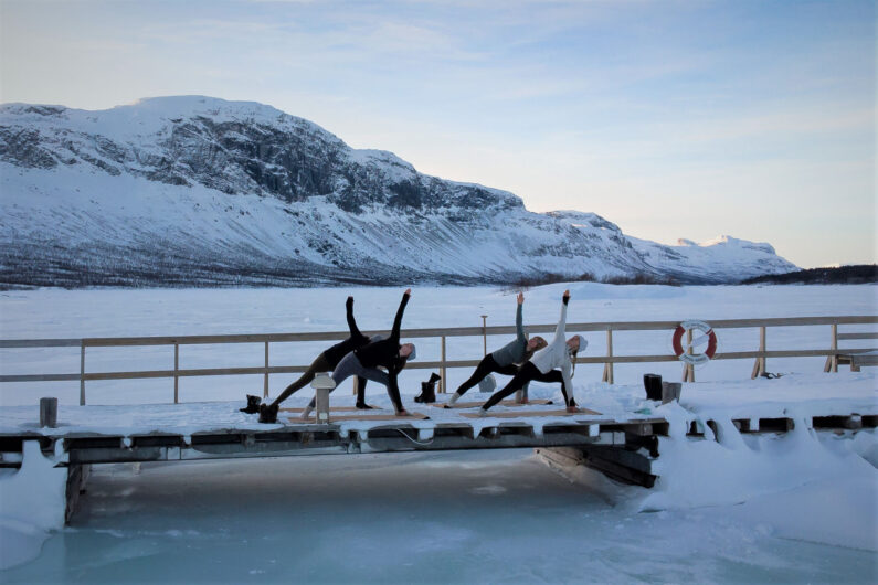En grupp utövar yoga på en brygga i vinterlandskap vid Saltoluokta och utsikt över Stora Sjöfallets berg.