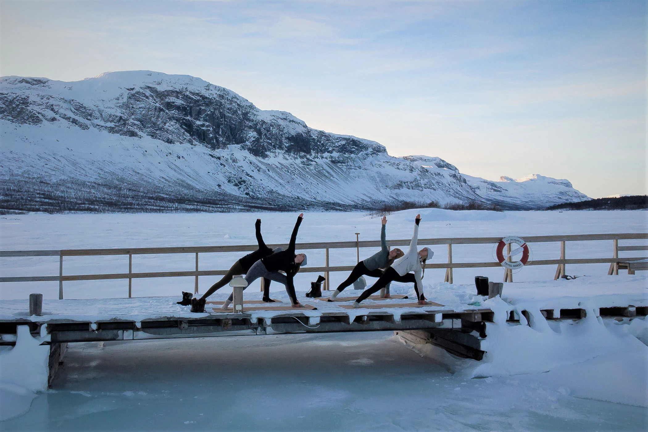 En grupp utövar yoga på en brygga i vinterlandskap vid Saltoluokta och utsikt över Stora Sjöfallets berg.