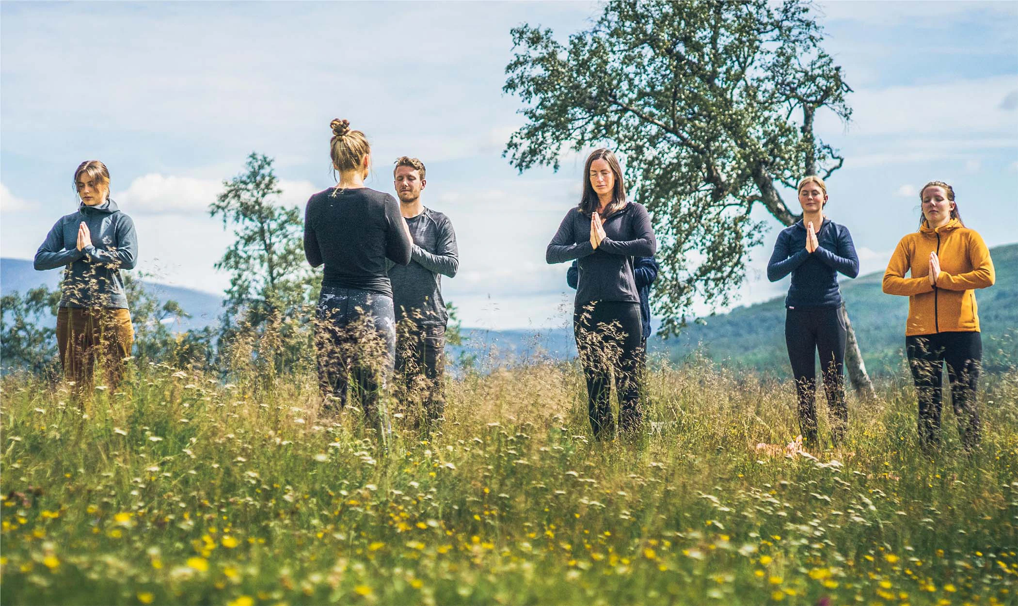 En grupp människor gör yoga utomhus på en blomsteräng med fjäll i bakgrunden.