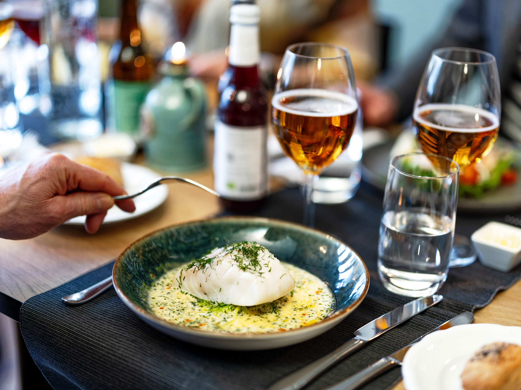 Ett uppdukat bord med ett par glas öl, en tallrik med fisk och en hand med en gaffel.