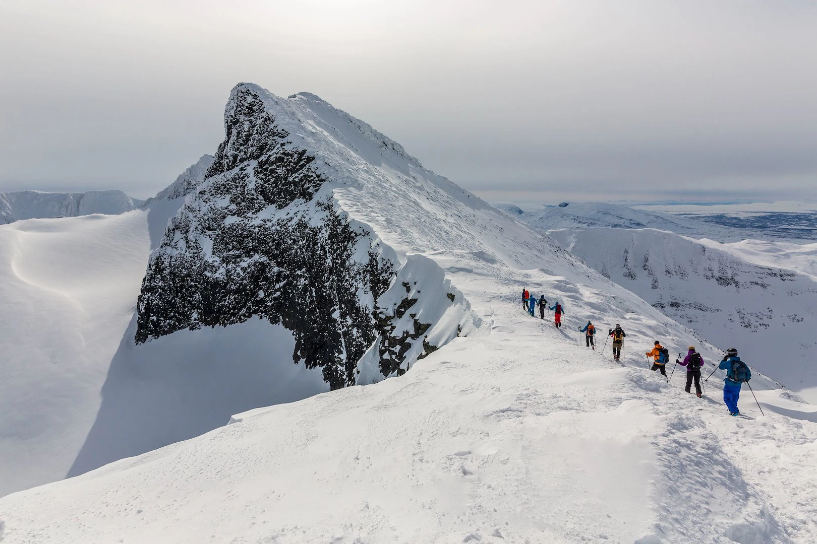 En grupp människor vandrar på en snötäckt fjällrygg med en dramatisk bergstopp i bakgrunden.