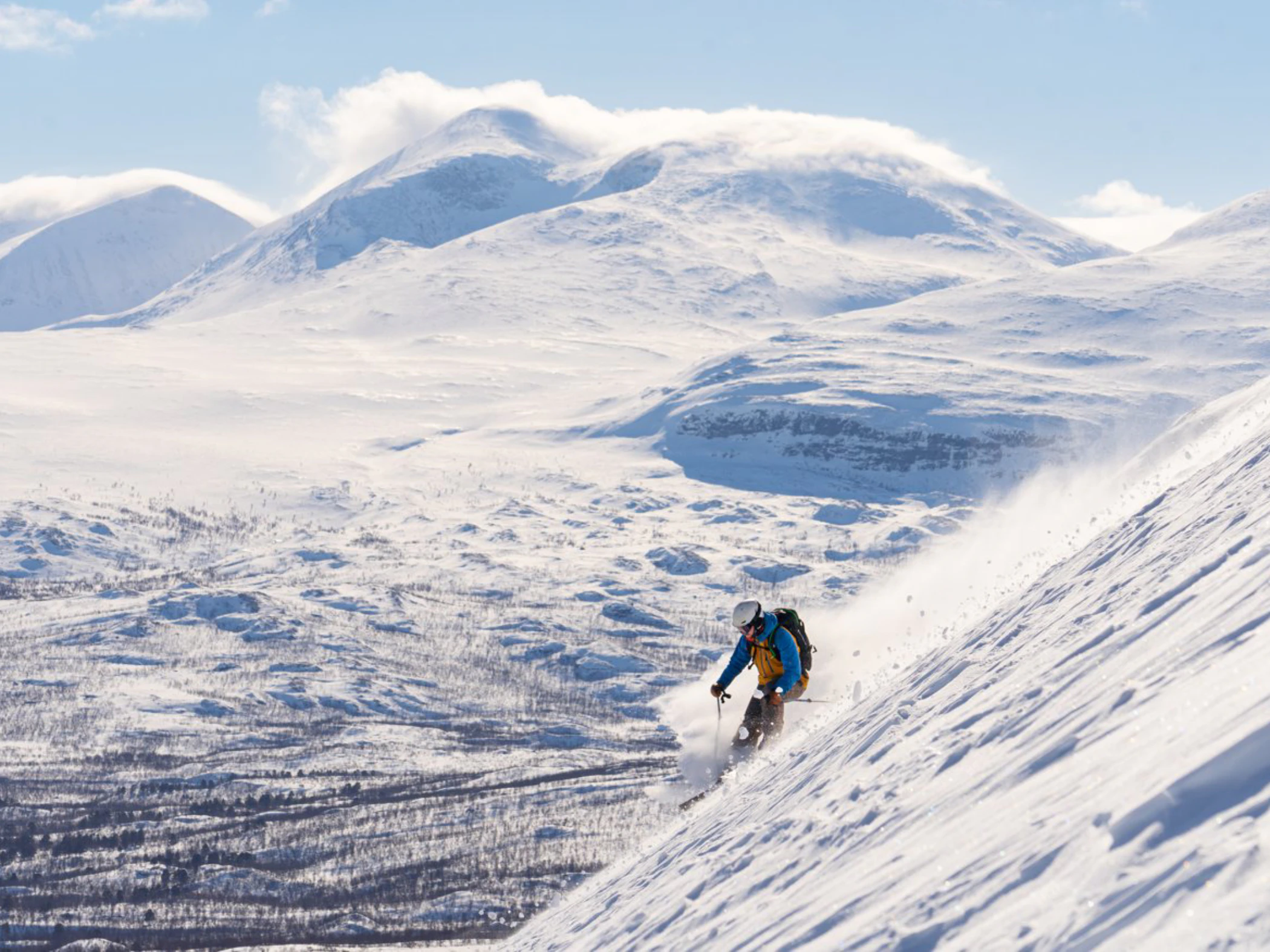Skidåkare som åker nedför brant fjällsluttning med snöklädda berg i bakgrunden.