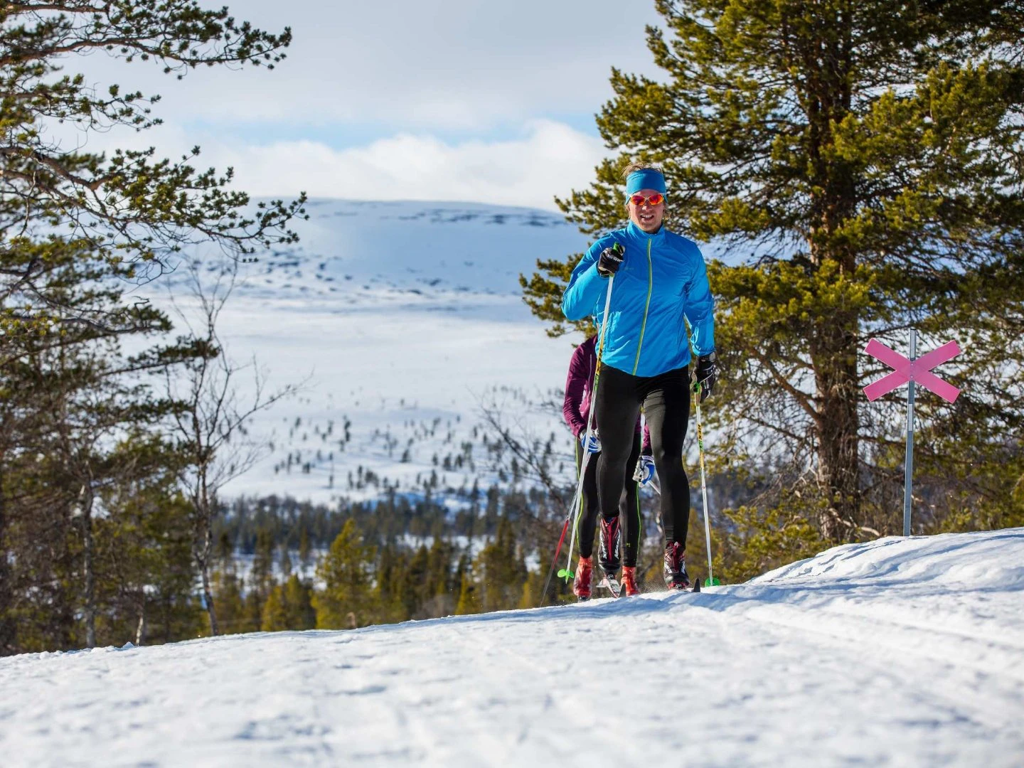Längdskidåkare i blå jacka som åker på markerad led i fjällskog med snöklädda berg i bakgrunden