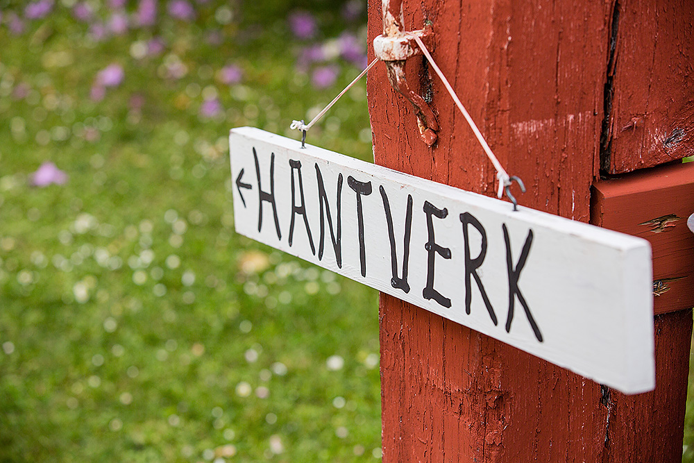 Handmålad träskylt med texten 'Hantverk' hänger på ett rött trästaket, med grönskande sommaräng i bakgrunden.