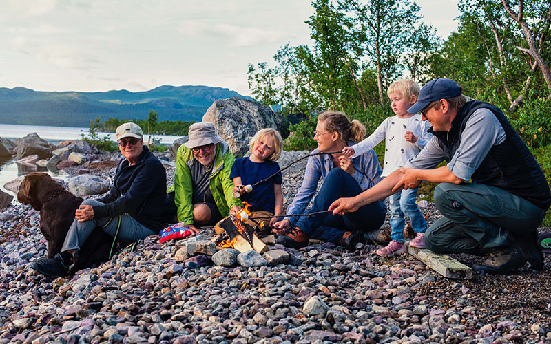 Familj med äldre, barn och hund grillar på en strand i fjällen.