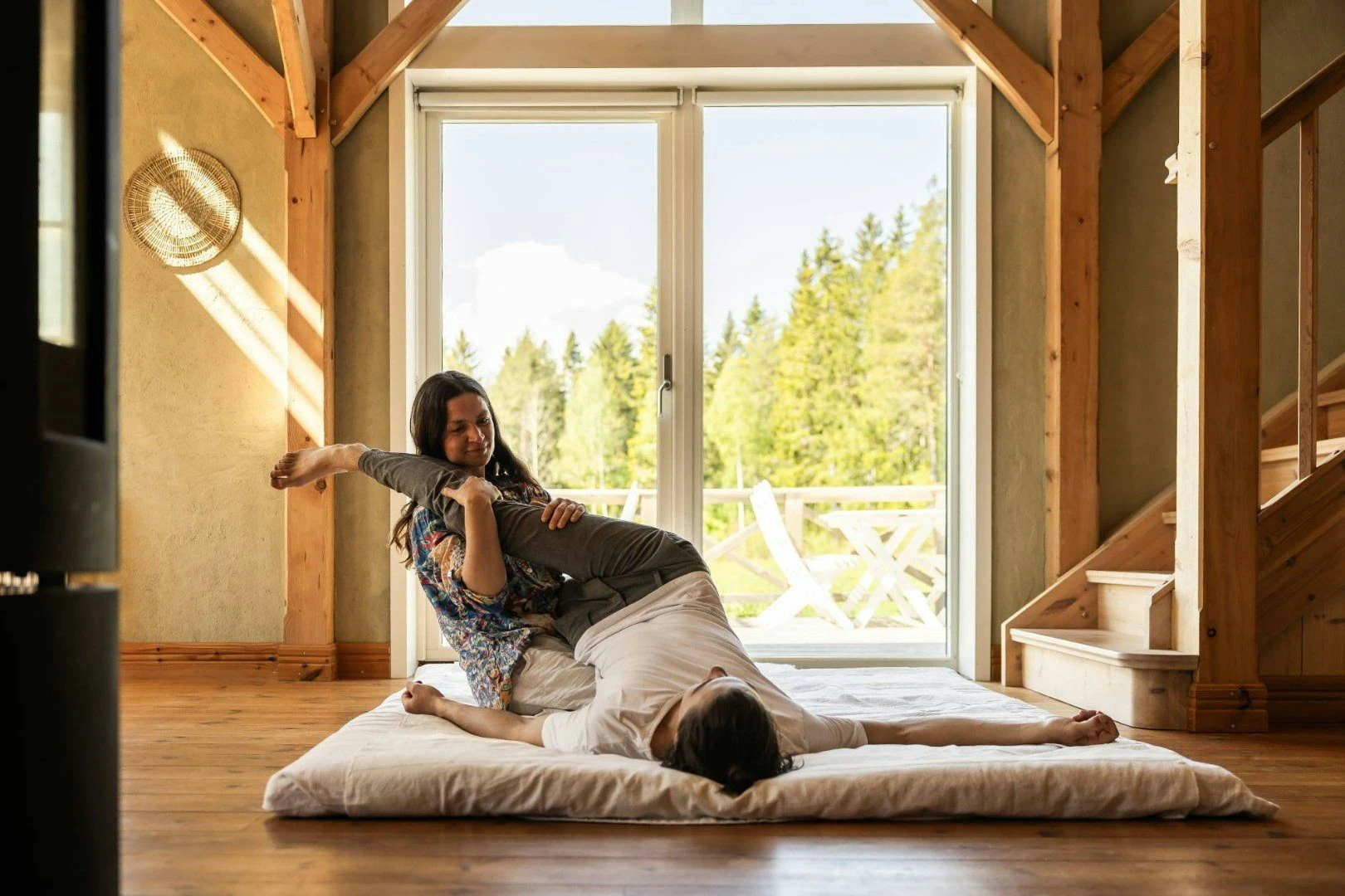 Person får yogamassage och assisterad stretchbehandling på en madrass i ett ljust rum med stora fönster.
