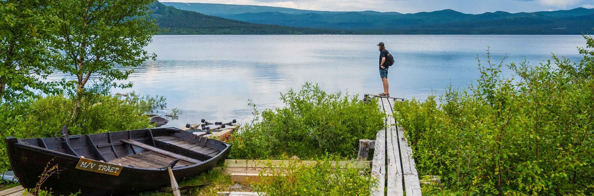 En person står på en smal träspång vid en sjö och blickar ut över det stilla vattnet, med fjäll i bakgrunden och en gammal roddbåt förtöjd i strandkanten.