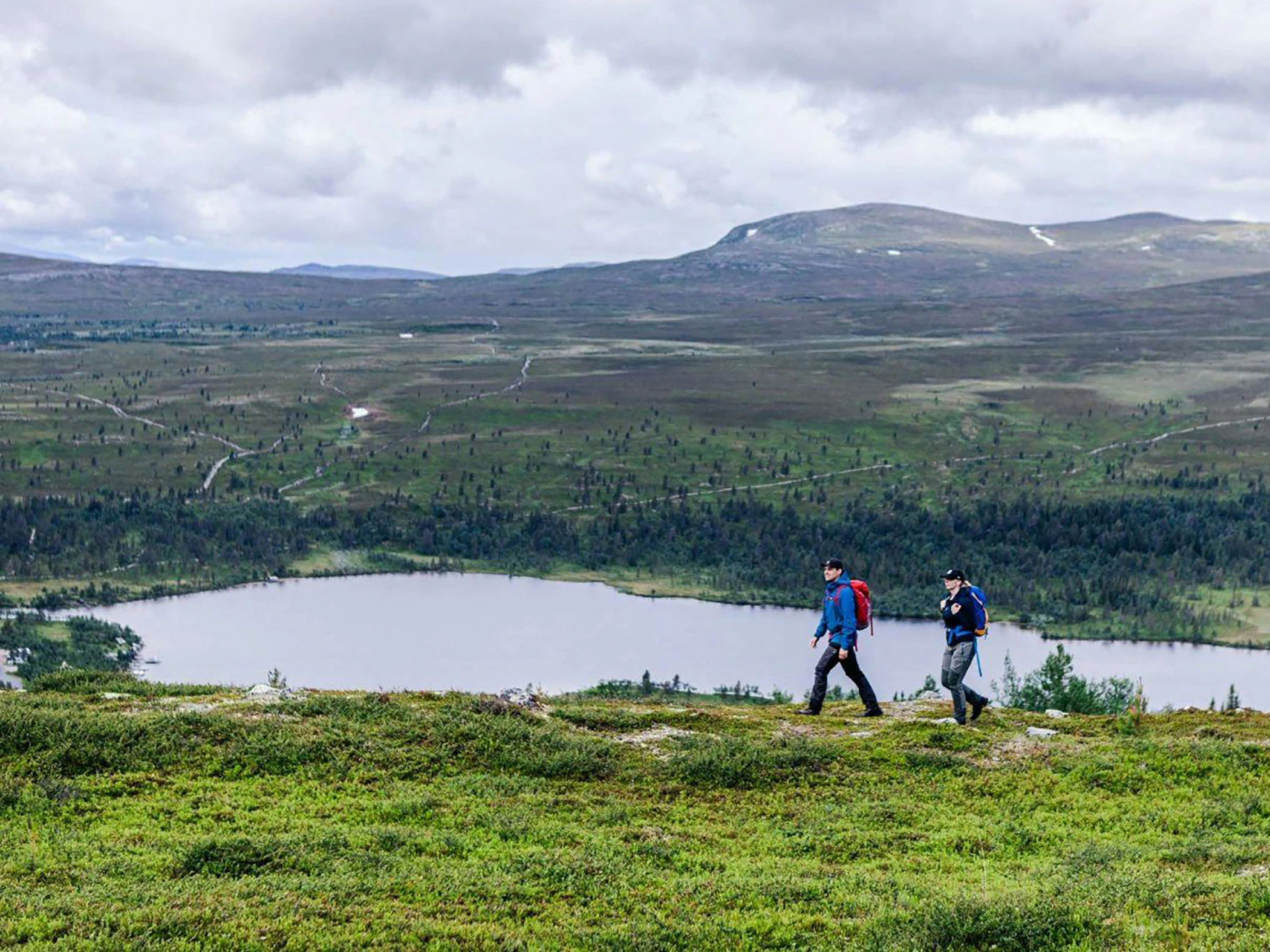 Två personer vandrar. I bakgrunden skymtar sjö och fjäll.