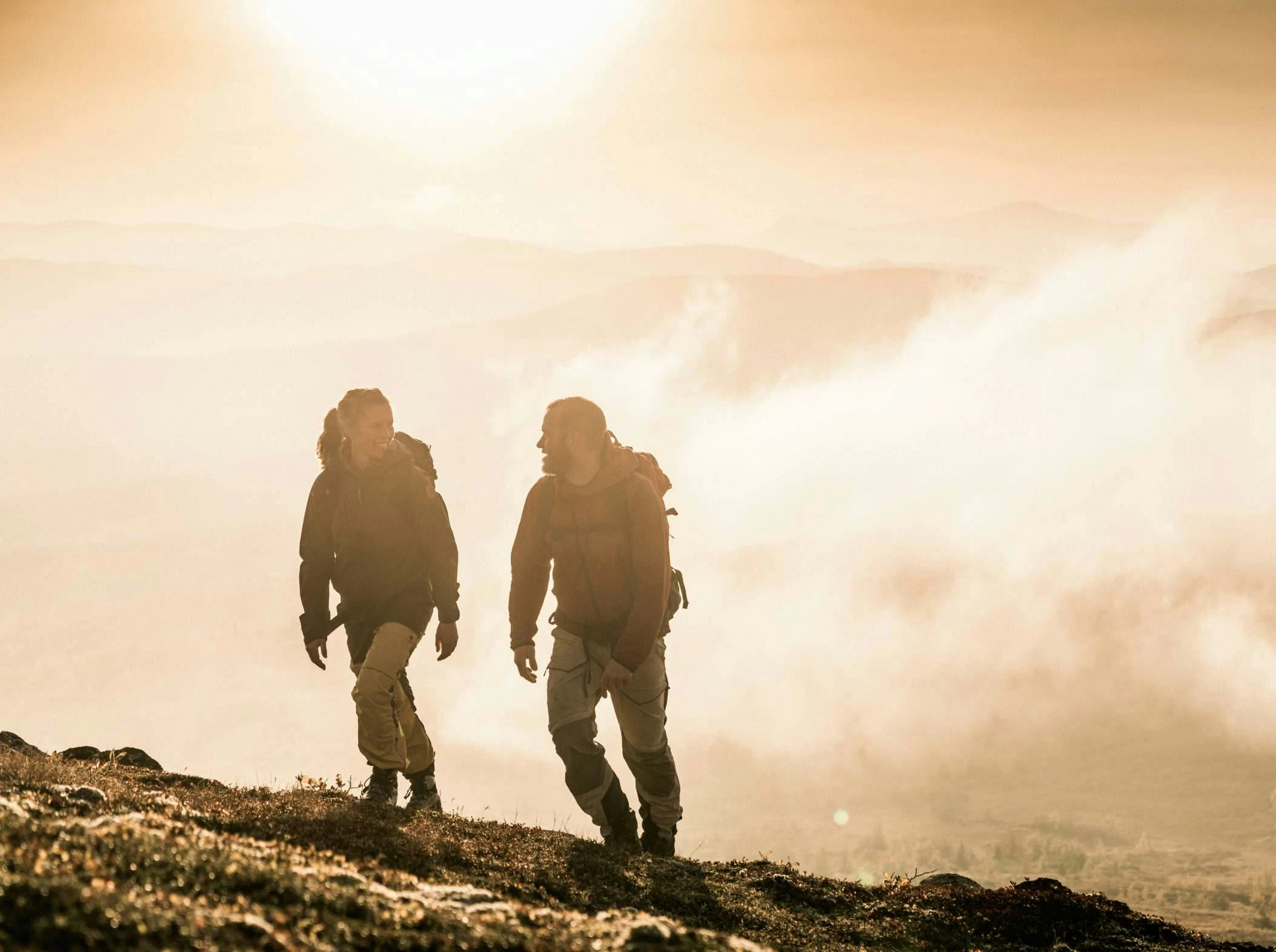 Två vandrare går uppför ett fjäll i gyllene ljus från soluppgång eller solnedgång, med dimma och fjäll i bakgrunden.