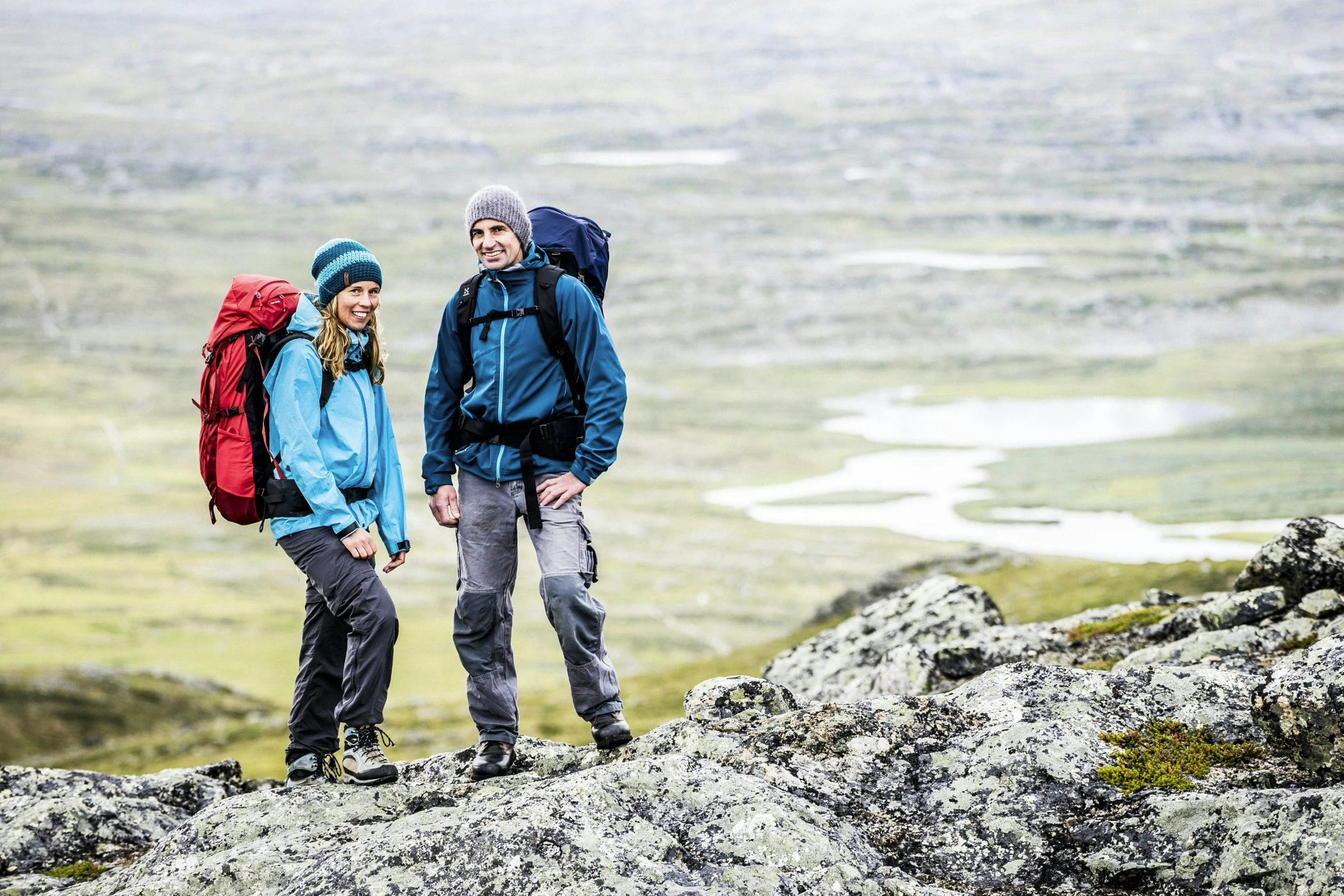 Två vandrare med ryggsäckar står på en stenig höjd med utsikt över ett fjällandskap.