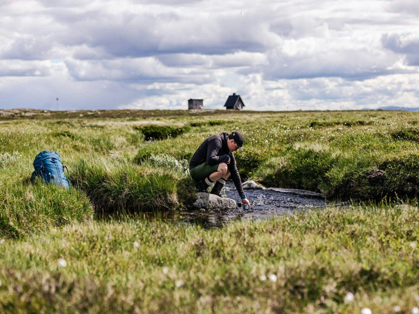 En person fyller en vattenflaska i en bäck, med en ryggsäck bredvid och fjällstugor i bakgrunden.