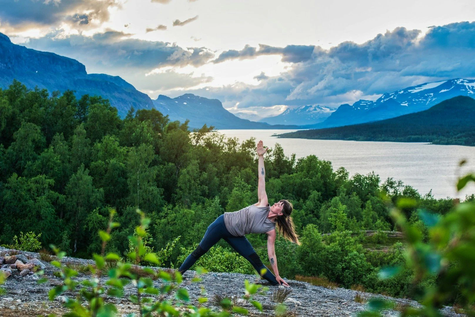 Person gör yoga i naturen med utsikt över fjäll, skog och sjö i solnedgången.