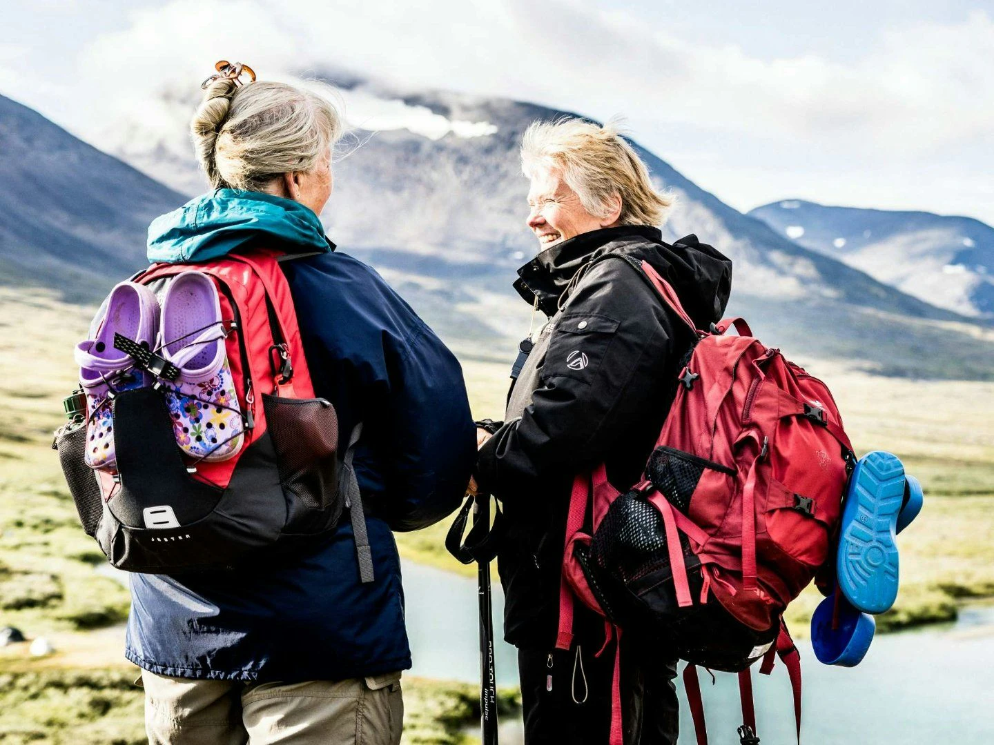 Två vandrare med ryggsäckar står i en fjällmiljö med gräs, en forsande å och berg i bakgrunden.