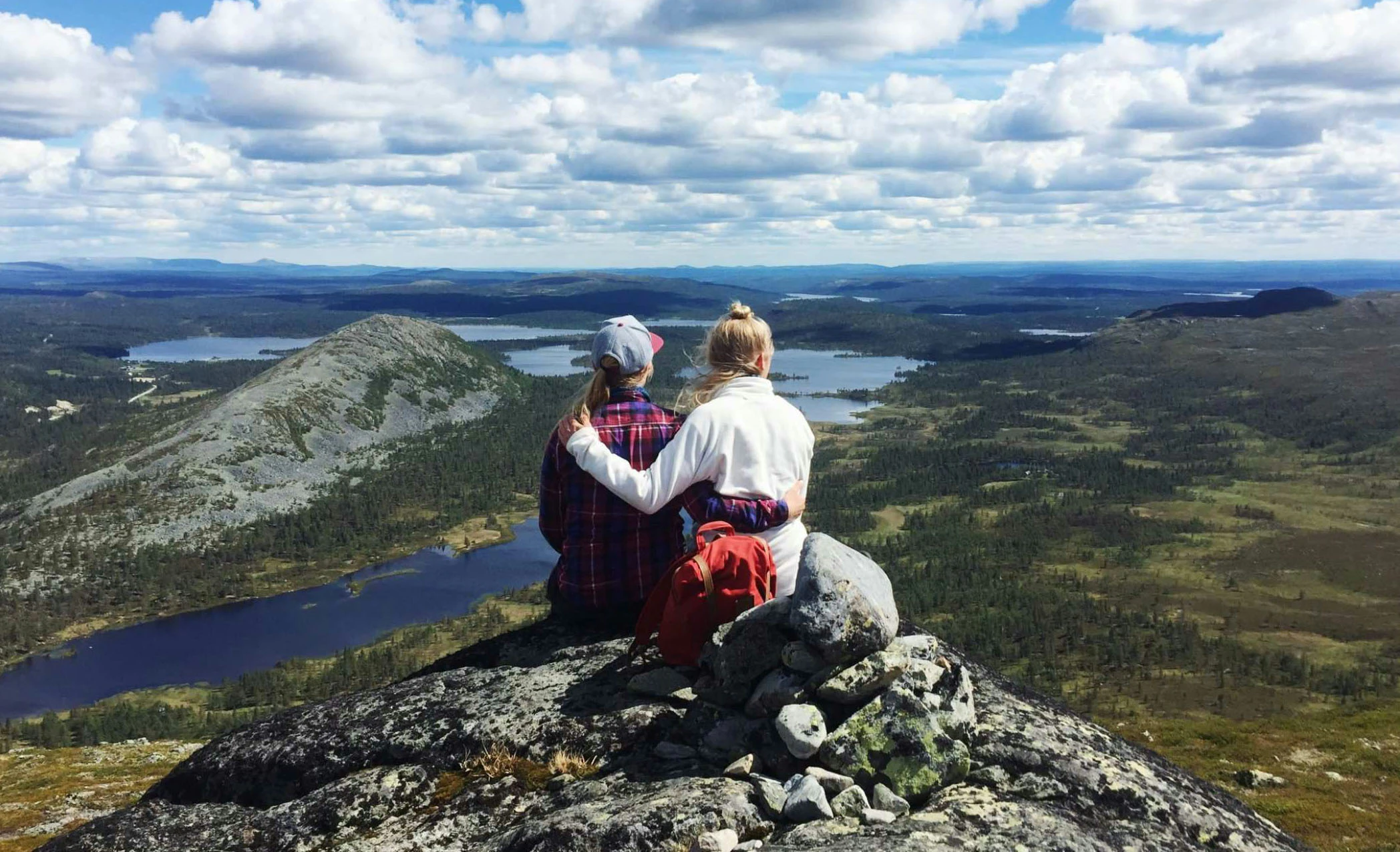 Två personer sitter på en bergstopp och blickar ut över ett vidsträckt landskap med sjöar, skogar och berg under en klarblå himmel med moln.