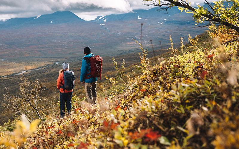 Två vandrare står på en höstfärgad fjällsluttning och blickar ut över ett vidsträckt landskap.